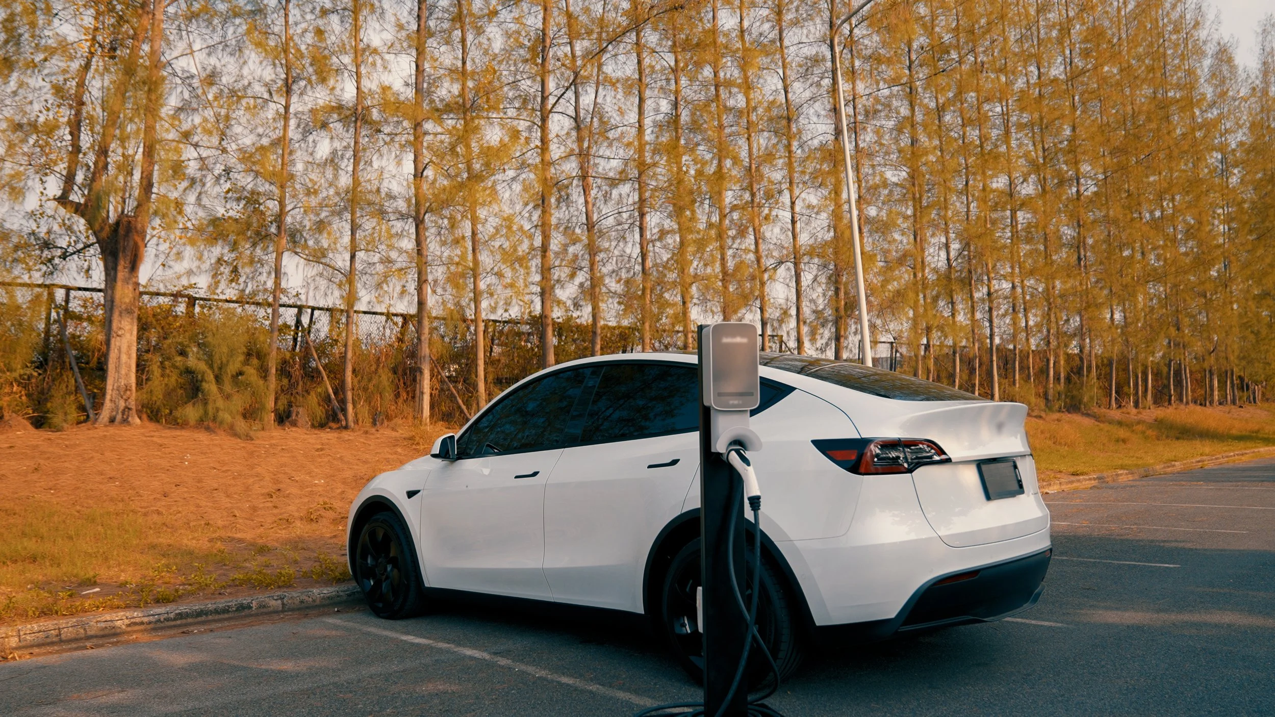 Voiture électrique blanche branchée à une borne de recharge dans un parking entouré d'arbres en automne.