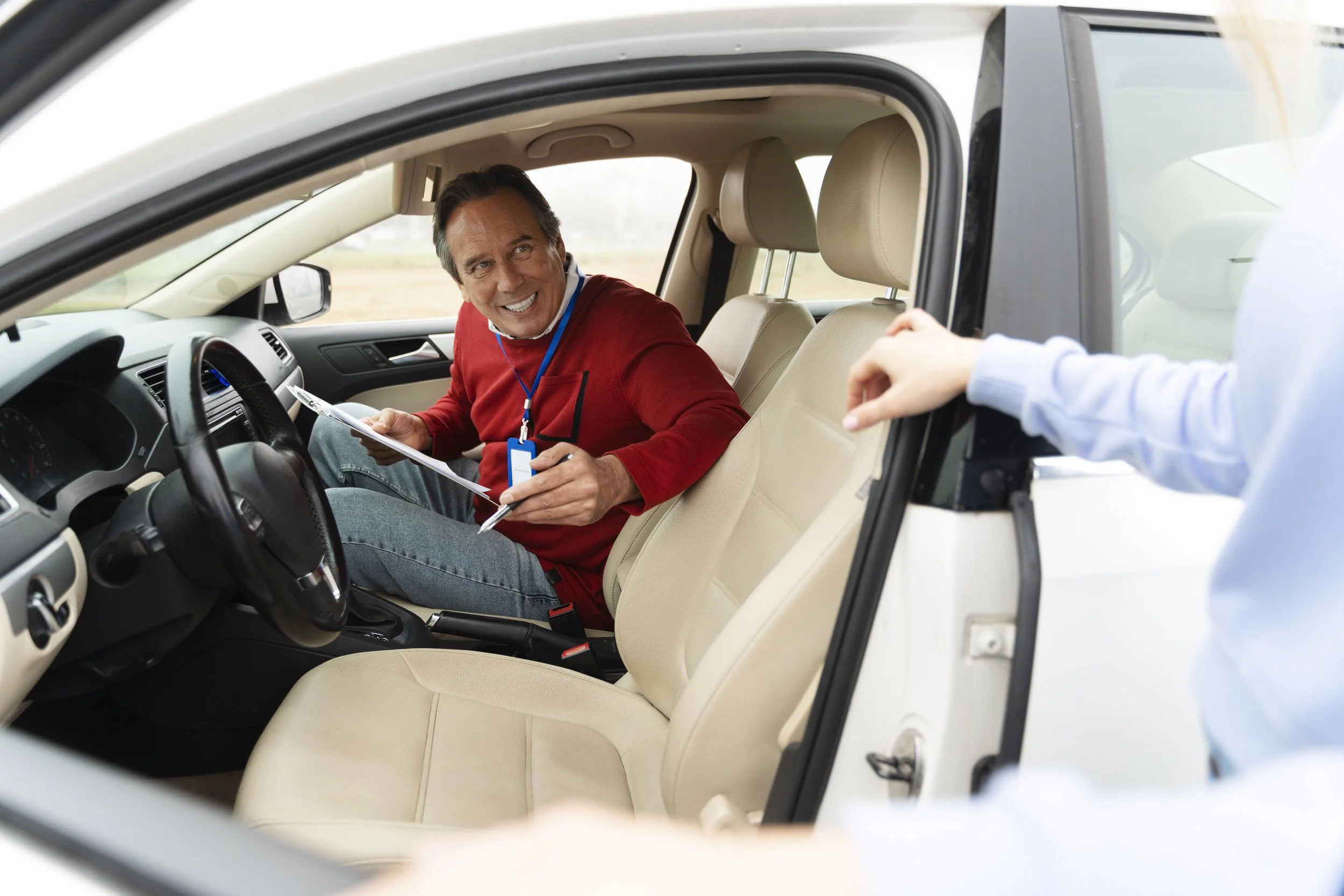 Un homme souriant est assis dans une voiture, parlant avec quelqu'un à l'extérieur, la main de cette personne est visible par la fenêtre.