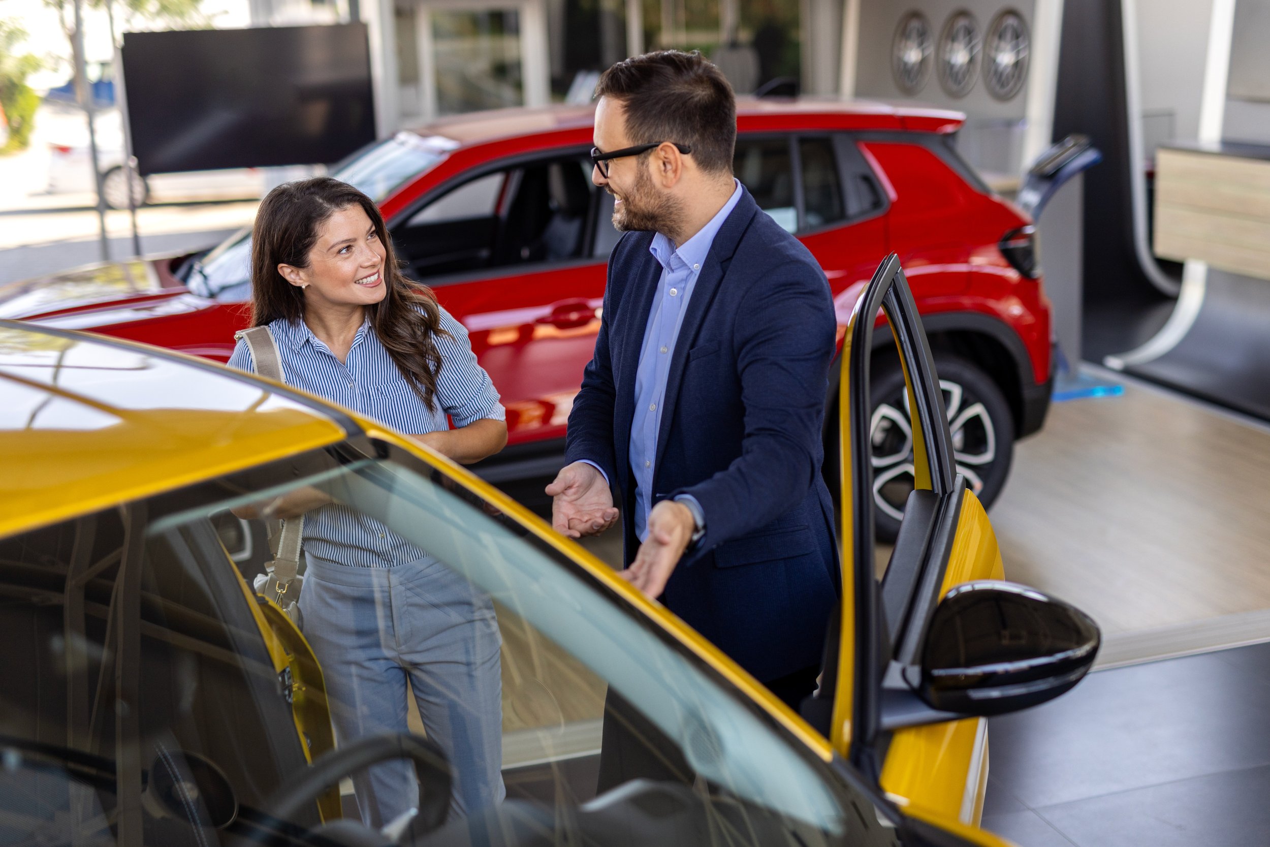 Un homme et une femme discutent devant une voiture jaune dans un concessionnaire automobile, avec d'autres voitures en arrière-plan.