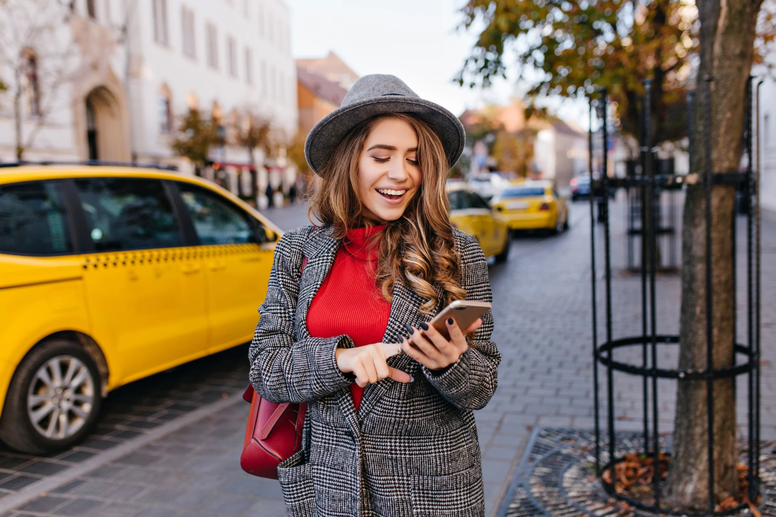Jeune femme souriante utilisant un smartphone dans une rue urbaine avec des taxis jaunes et des arbres en automne.