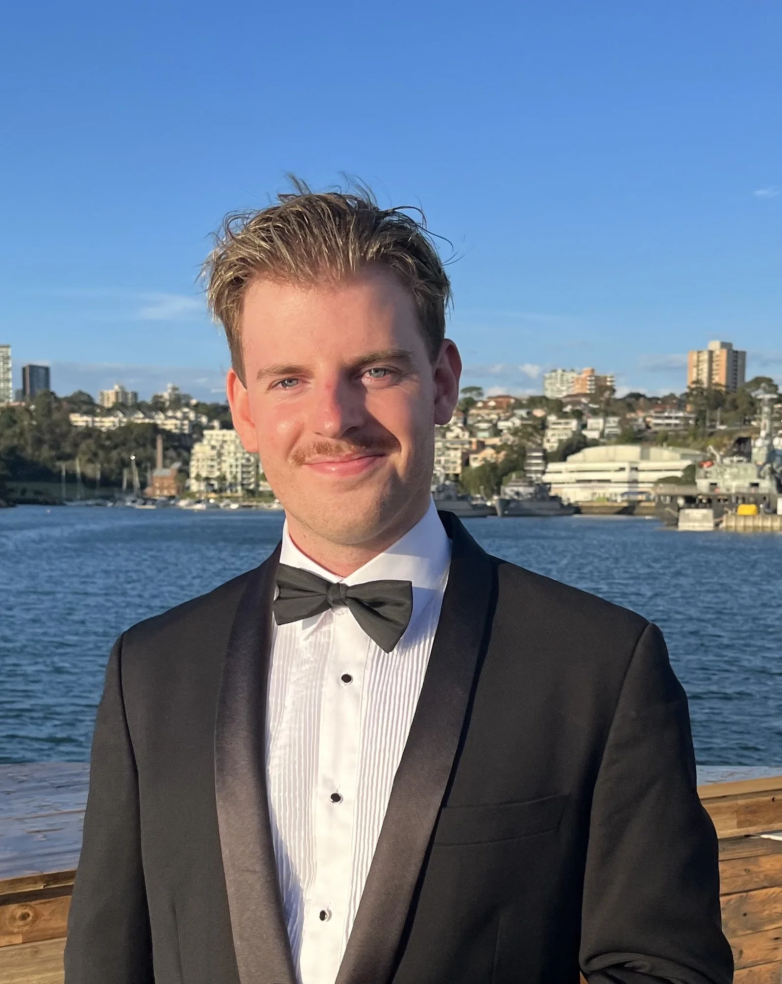 A young man in a tuxedo standing outdoors near the water with a cityscape in the background, smiling at the camera.