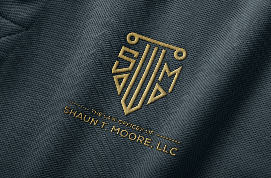 Close-up of a fabric with a gold embossed logo and text. The logo is a shield featuring the letters "S" and "M". The text reads 'The Law Offices of Shaun T. Moore, LLC'.
