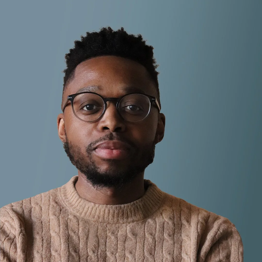 Close-up portrait of a man with glasses, a beard, and short curly hair, wearing a beige knitted sweater, against a plain blue background.