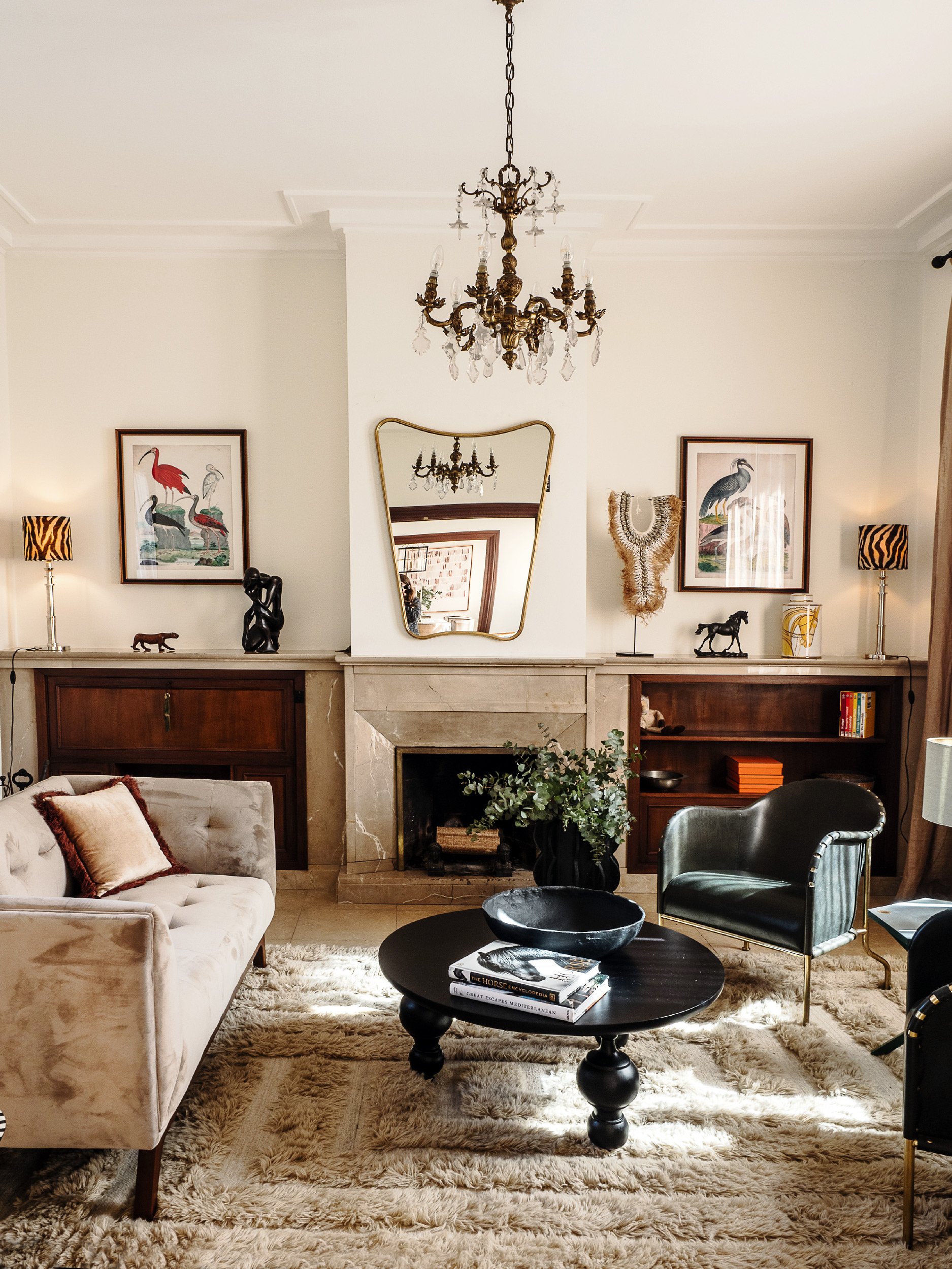 A living room with a chandelier, two framed bird artworks, a mirror, a fireplace, a beige sofa with cushions, a round black coffee table with books and a bowl, a black armchair, and decorative items on the shelves.