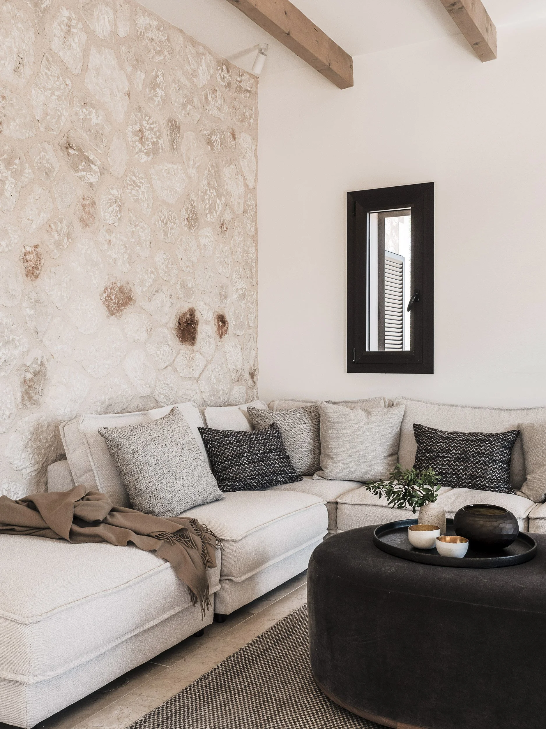 A cozy living room with a white sectional sofa decorated with black, white, and gray pillows. There is a tan throw blanket on the sofa. A dark round ottoman serves as a coffee table with black and white bowls on top. The room has a textured stone wal