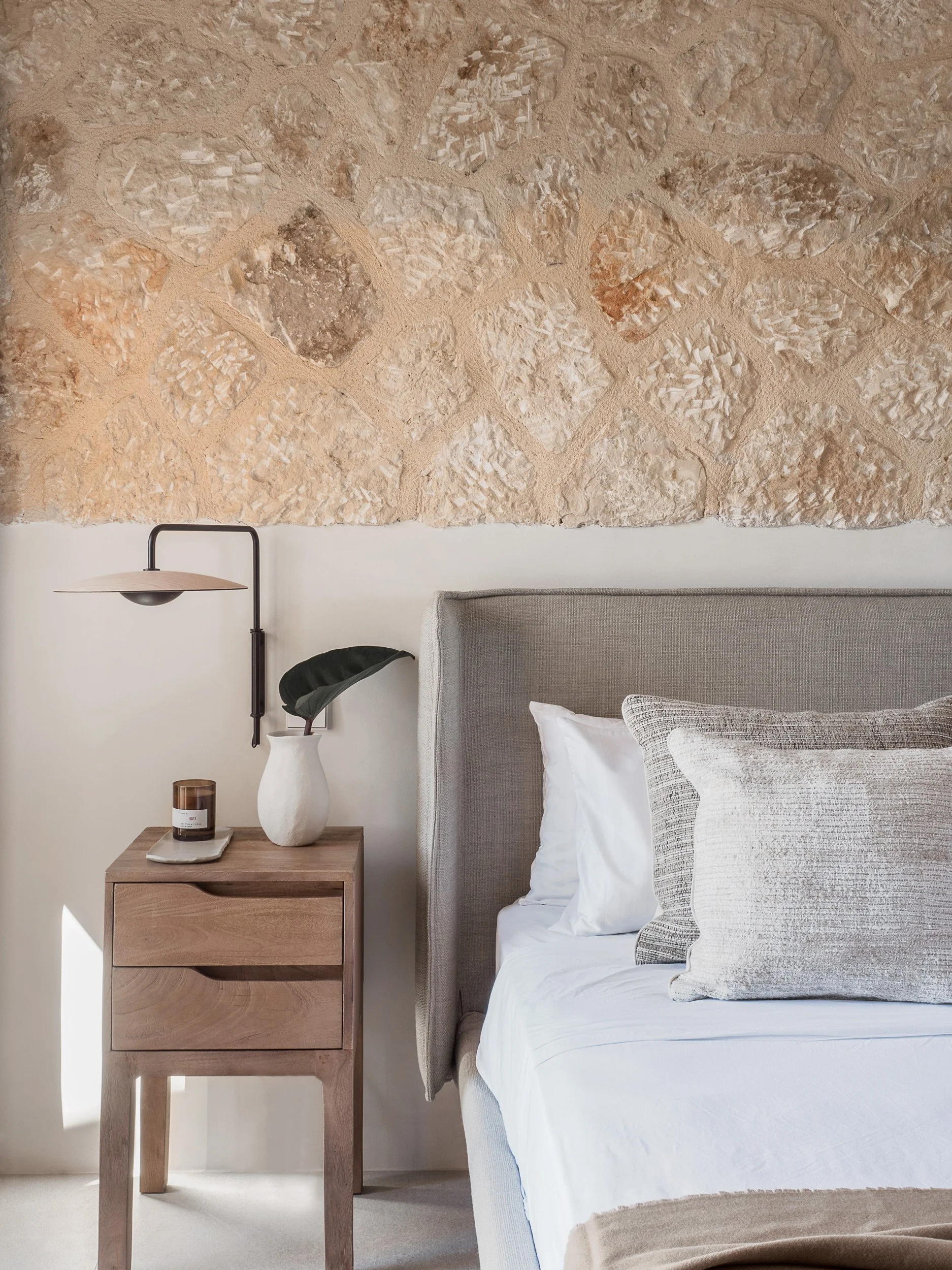 A bedroom with a stone wall above a bed with a gray headboard, white bedding, and patterned pillows. Next to the bed, there is a wooden nightstand with a white vase, a candle, and a tray. A black wall-mounted lamp is above the nightstand, and a leafy