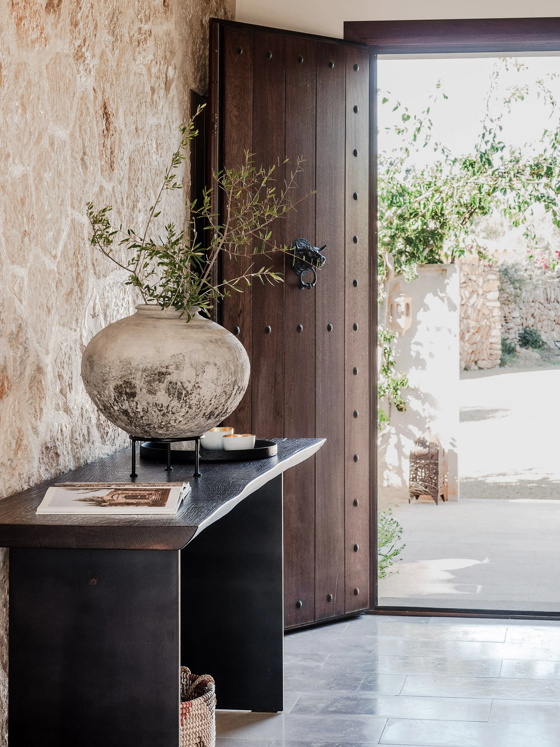 Open wooden door leading into a bright, outdoor space with stone and greenery, decorated with a large beige vase with greenery and a tray with small white bowls on a black table inside.