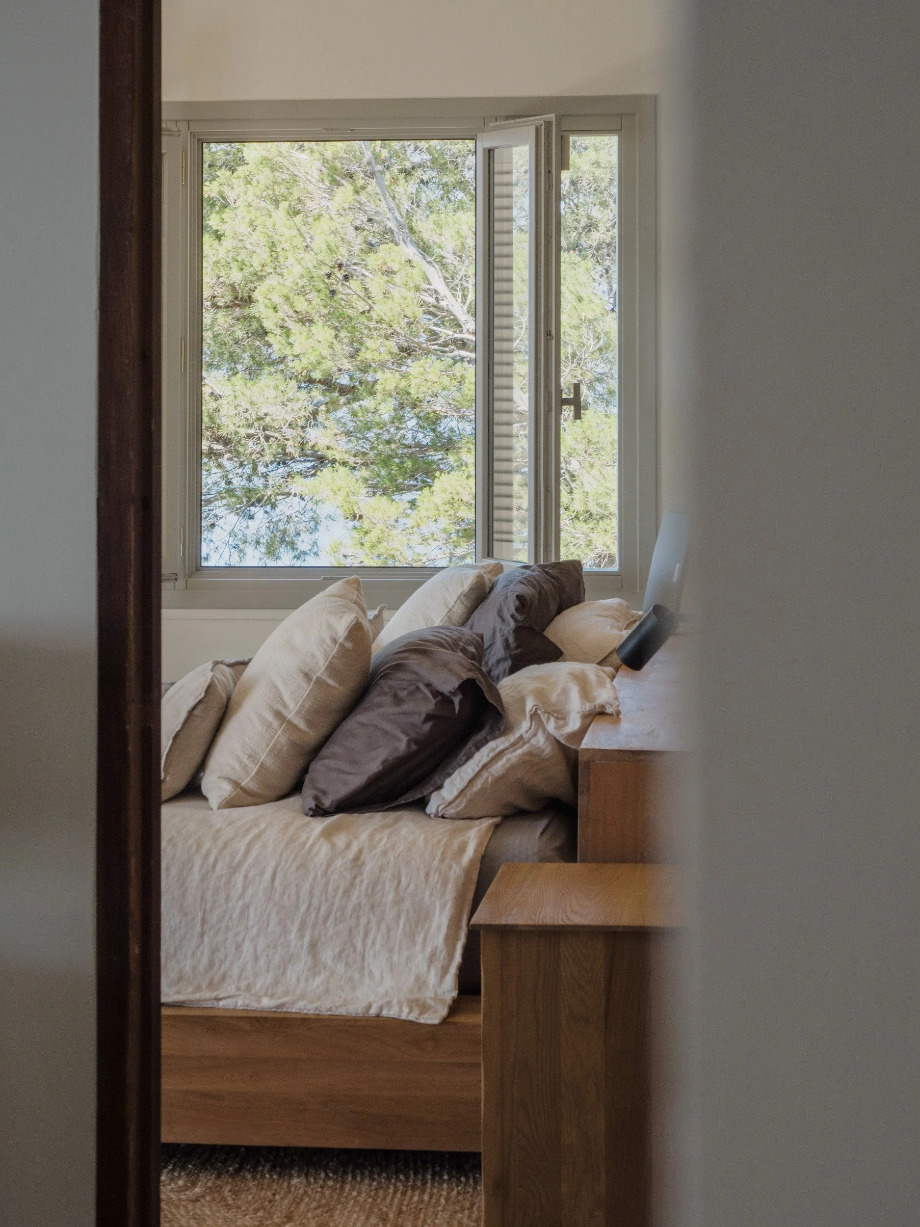 View of a bed with beige and brown pillows next to a window with trees outside, seen from a doorway in a bedroom.