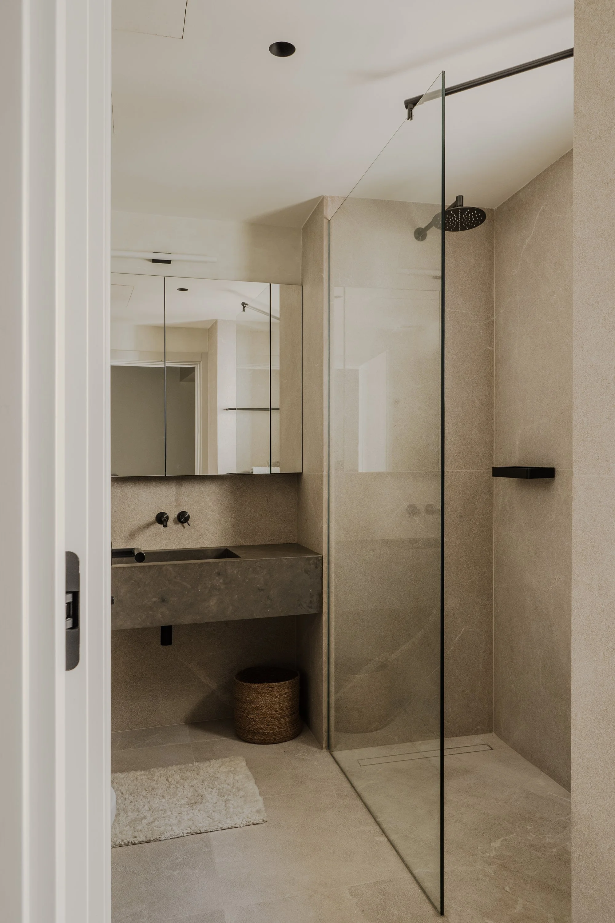 Modern bathroom with walk-in shower, beige tiles, and a large mirror. The shower has a black rain showerhead and a black shelf. There is a woven basket on the floor near a white plush rug.