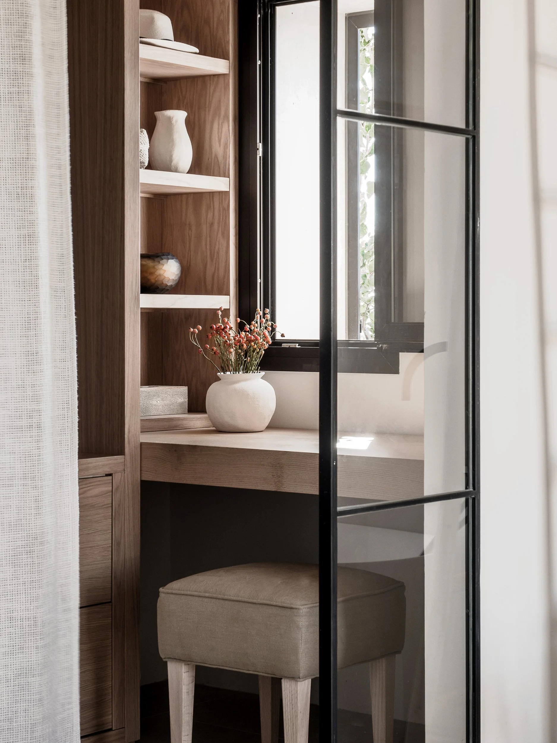 A small beige upholstered stool with wooden legs is positioned in a nook with a wooden shelf and a white vase with pinkish-red flowers. There's a window with black framing to the side, allowing natural light to illuminate the space.