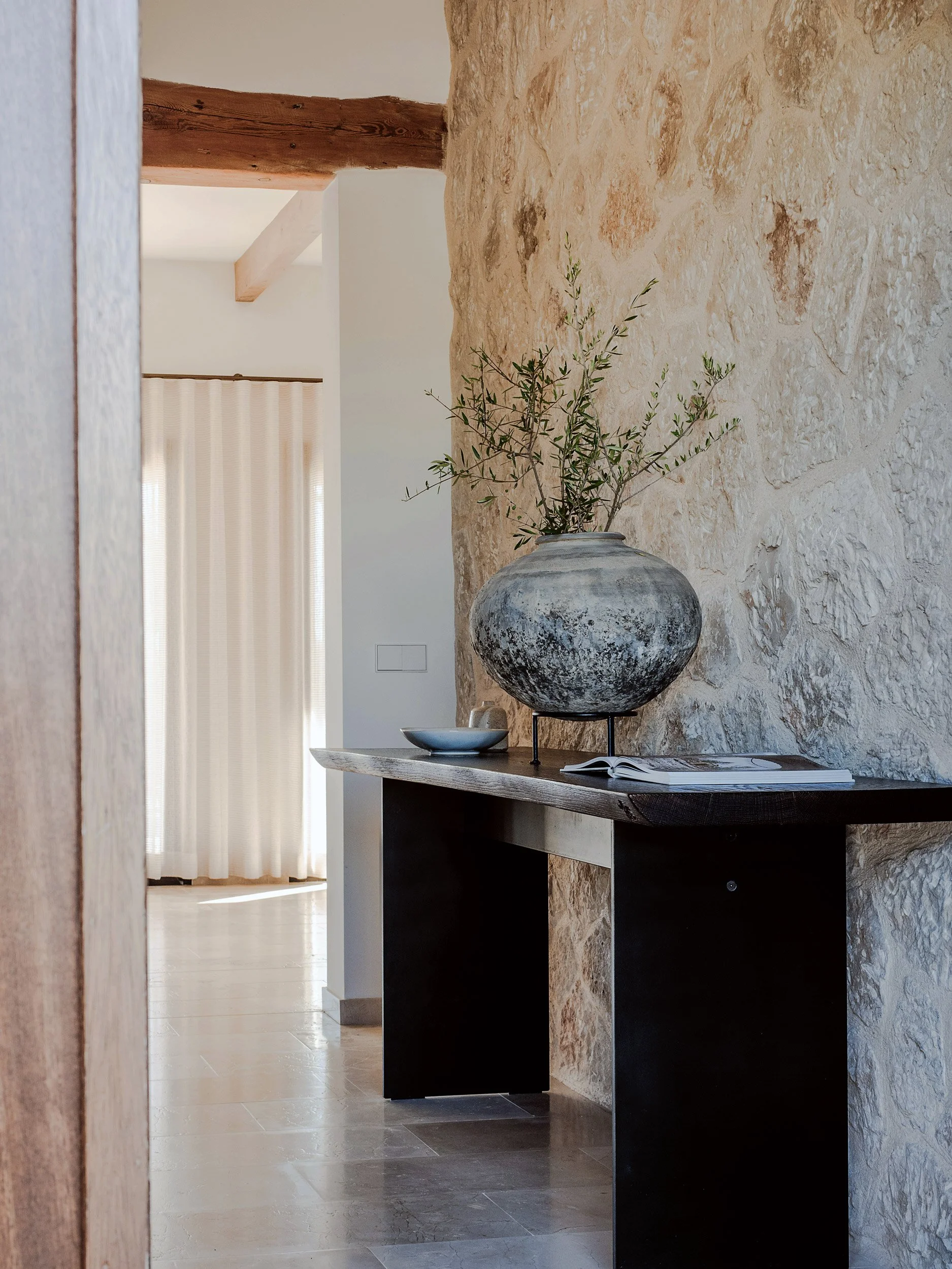 Interior view of a rustic room with a stone accent wall, featuring a dark wooden console table with a large, weathered ceramic vase holding green branches, and an open book resting on the table.
