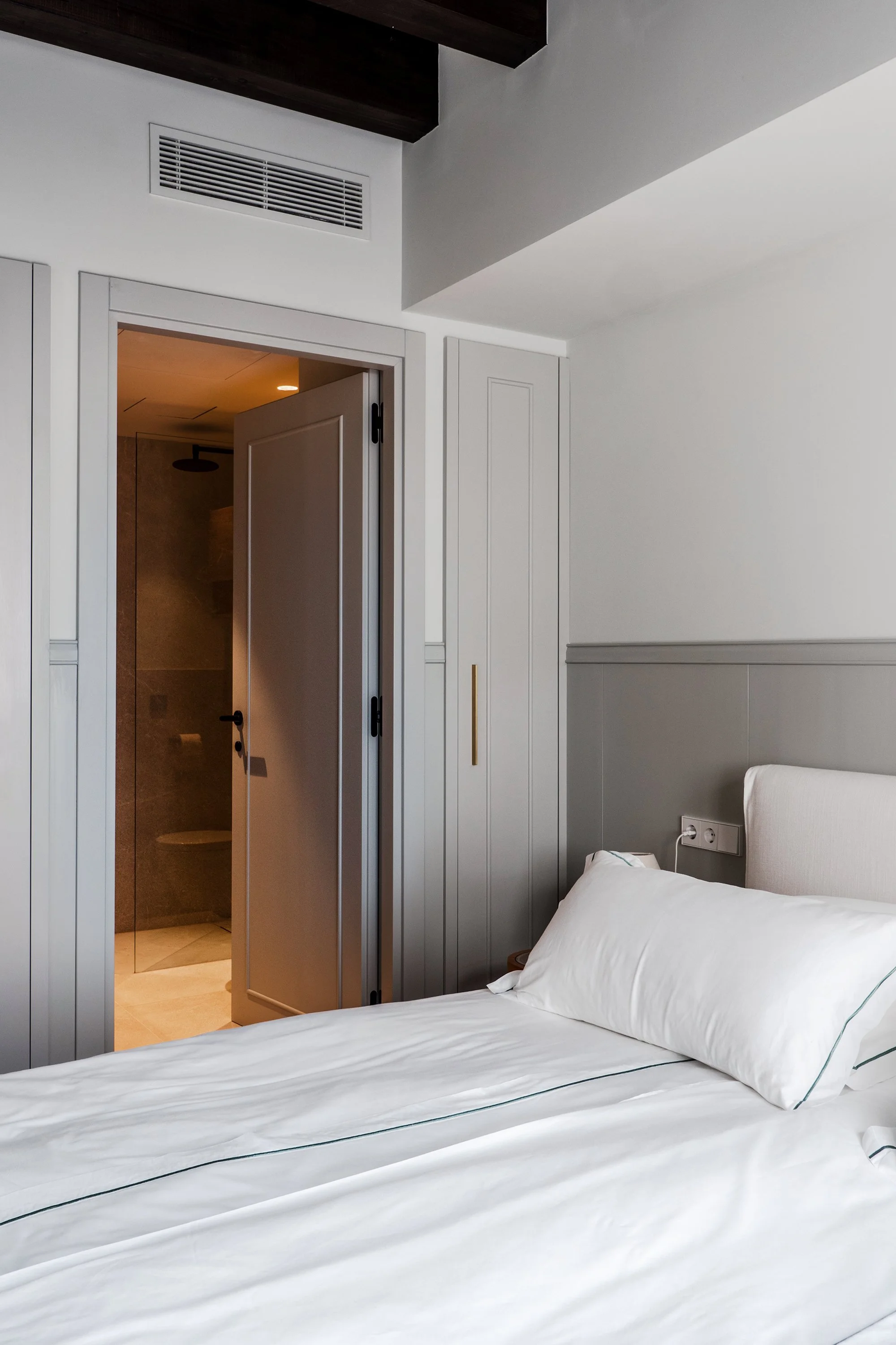 A bedroom with a white bed and pillow, visible electrical outlet, and a doorway leading to a bathroom with a shower and a hanging showerhead.