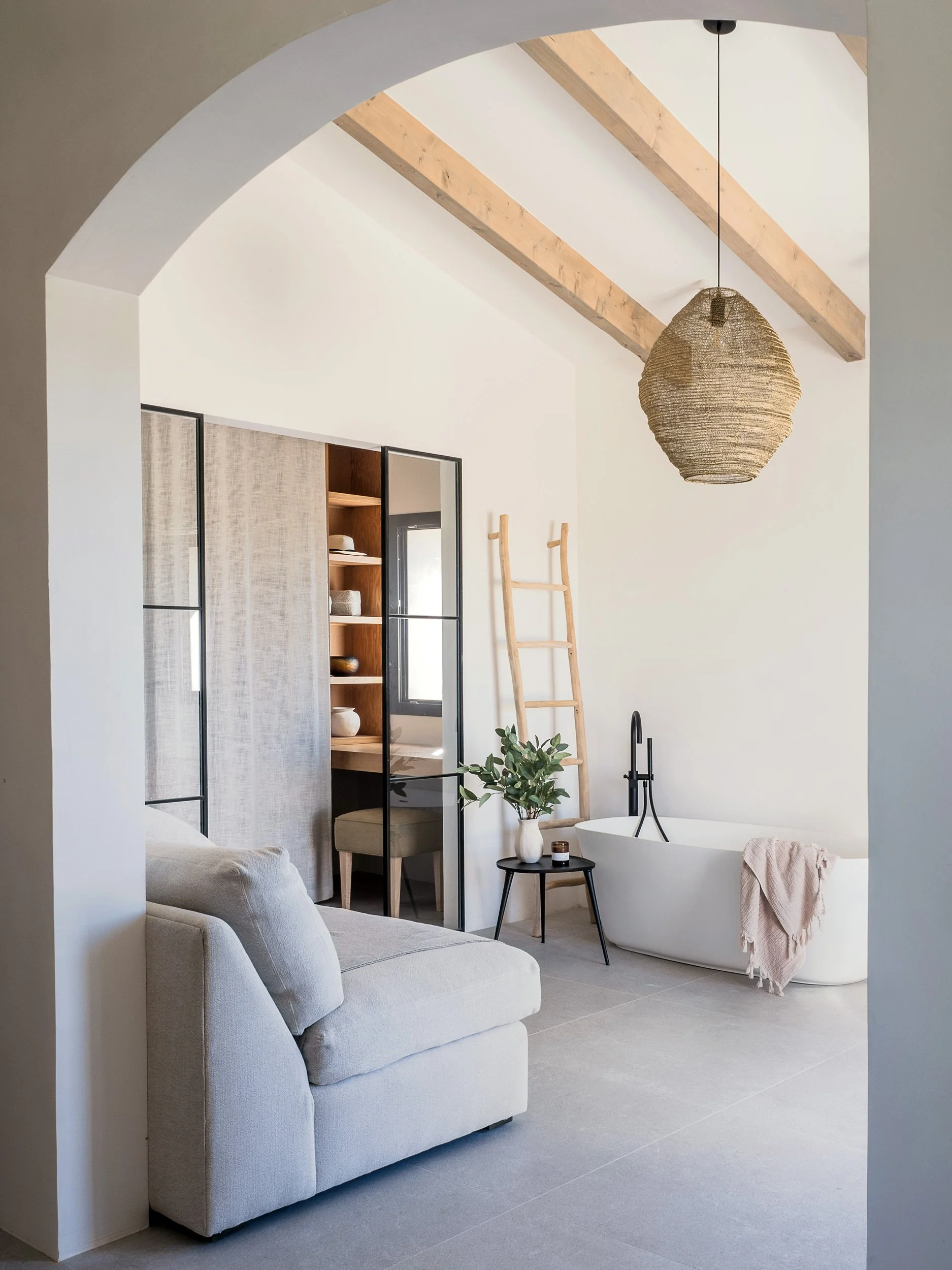 A minimalist bathroom with a freestanding bathtub, a small black side table with a potted plant and candle, a ladder, and a ceiling light fixture, with wood beams and an arched doorway.