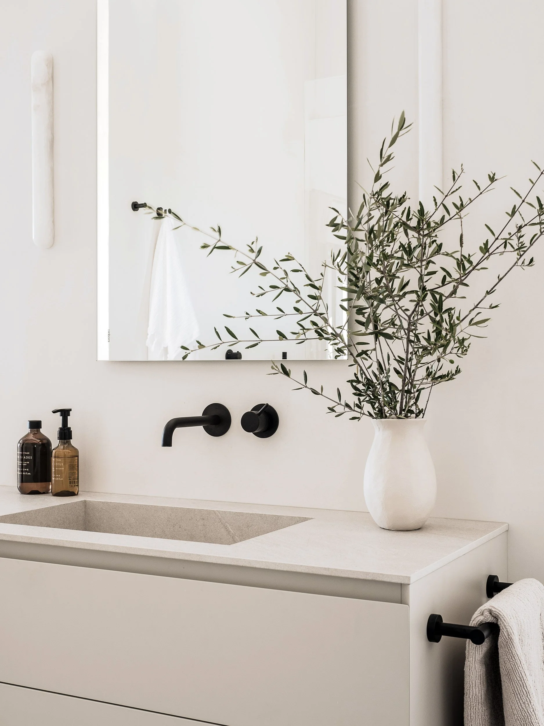 Minimalist bathroom countertop with a white sink, black wall-mounted faucet, two amber bottles, a large white vase with greenery, and a mirror reflecting a white towel and black hook.