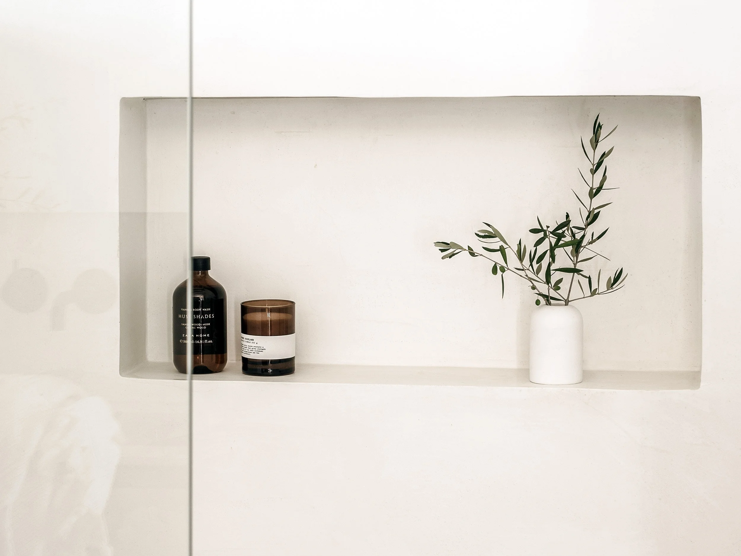 A white shelf with two brown bottles, a brown candle, and a white vase with green leafy branches against a white wall.