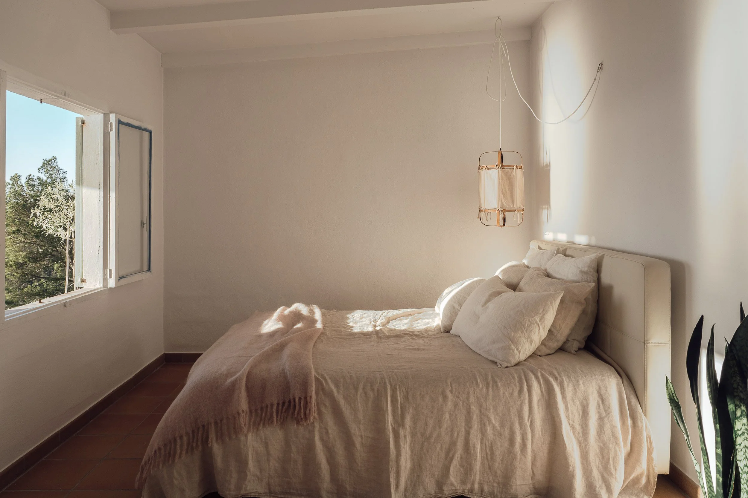 A bedroom with a bed covered in beige bedding and pillows. An open window on the left lets in natural light. A hanging lamp is suspended from the ceiling near the head of the bed. There is a plant with tall leaves on the right side of the image.