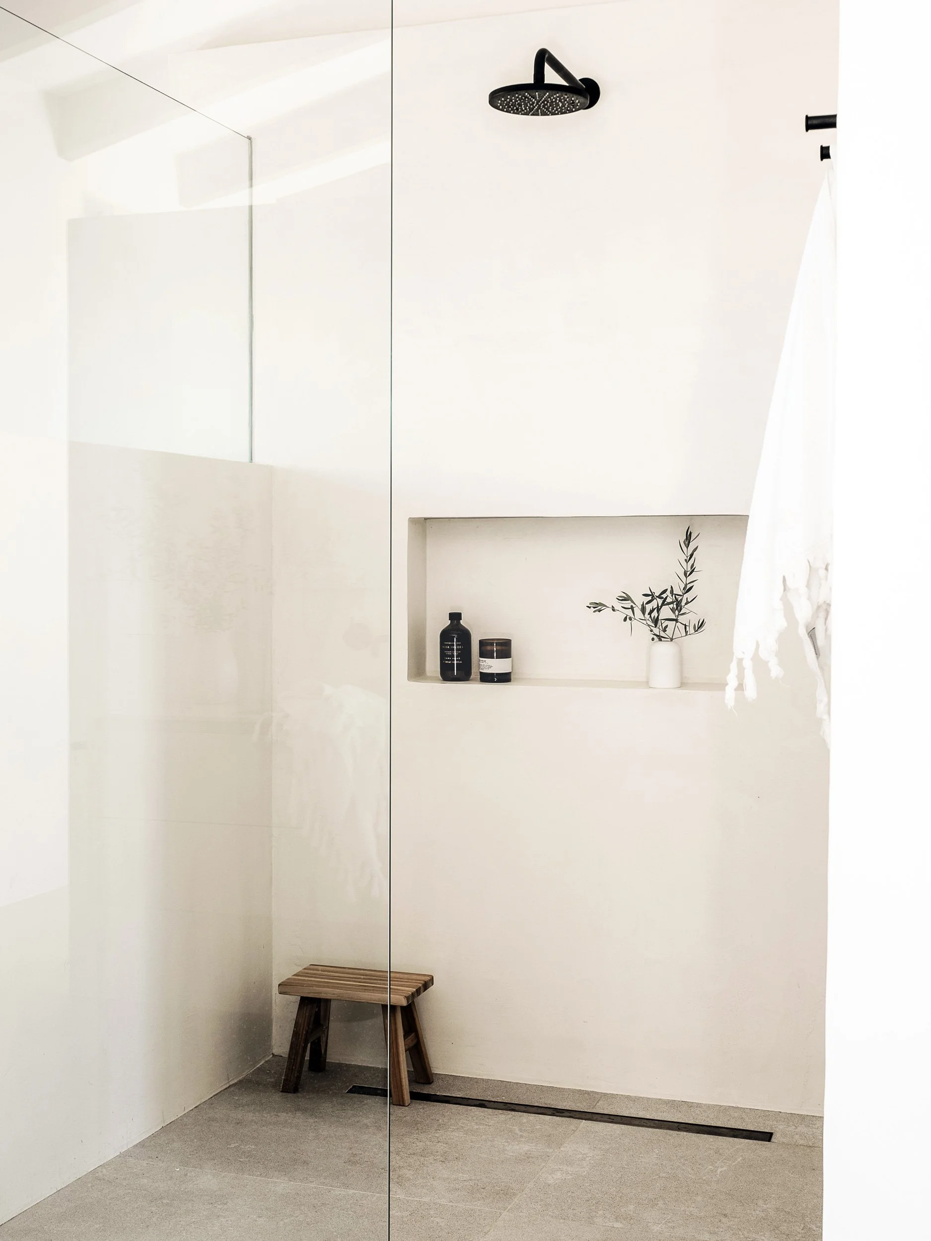Minimalist shower space with a black rain shower head on white wall, built-in niche containing black bottles and a small potted plant, with a wooden stool on beige tiled floor.