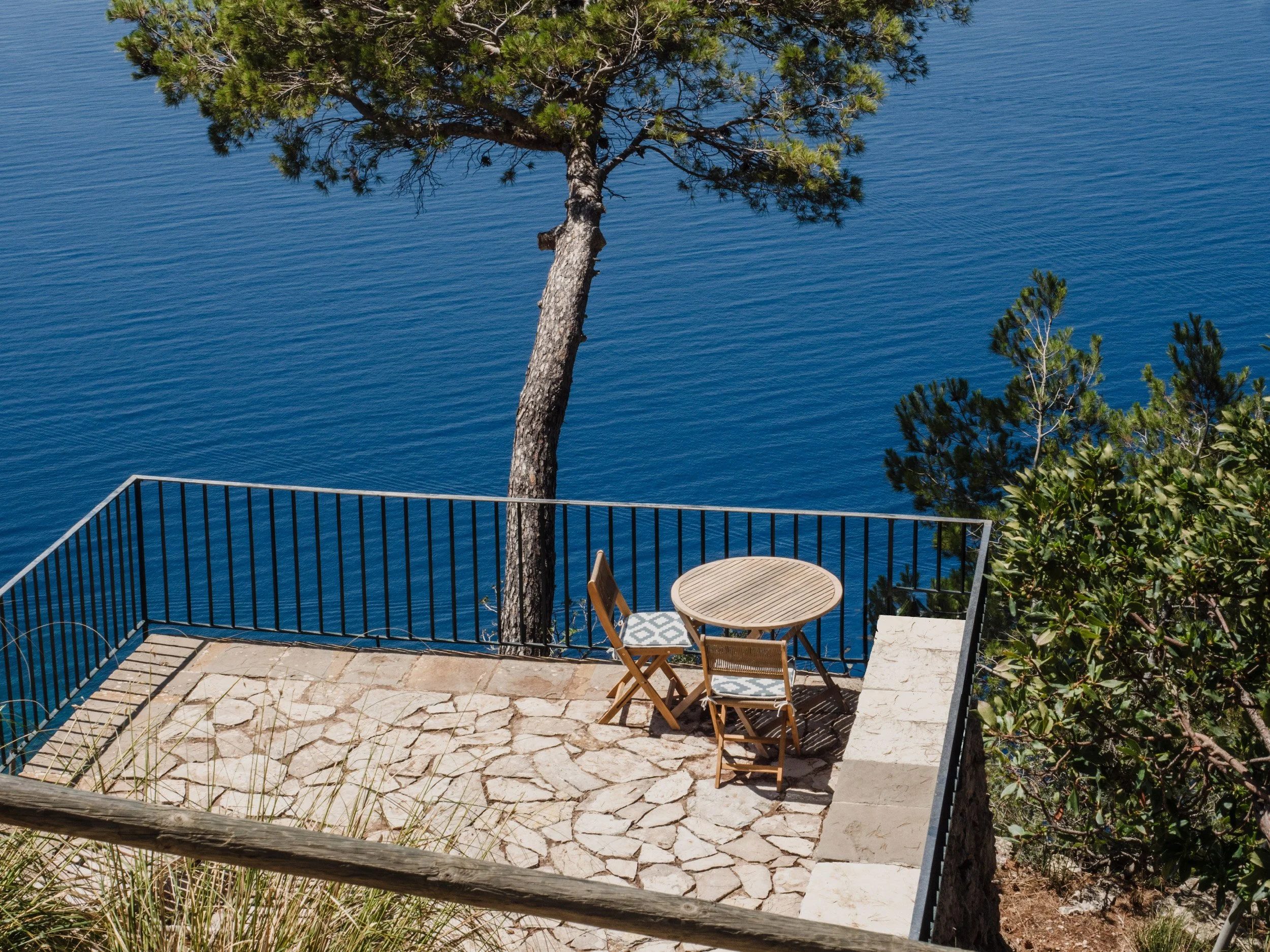 An outdoor patio area with a small round table and two chairs, overlooking a body of water with trees nearby. The patio is enclosed by a black metal fence, and there is greenery and trees surrounding the area.
