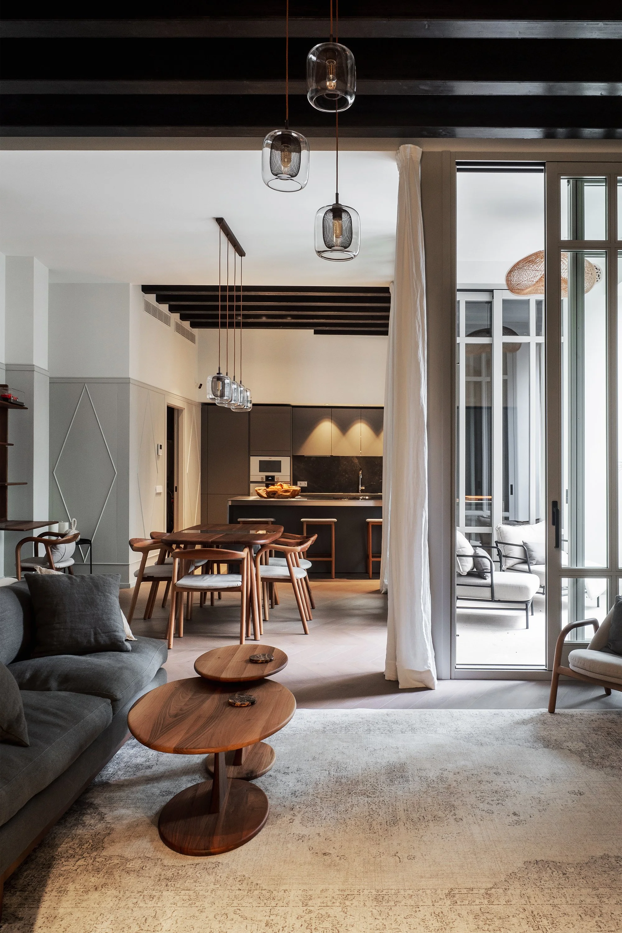 Living room with a grey sofa, wooden coffee tables, and a large cream-colored rug, adjacent to an open kitchen with a dining table and hanging pendant lights, with large windows and a door leading outside.