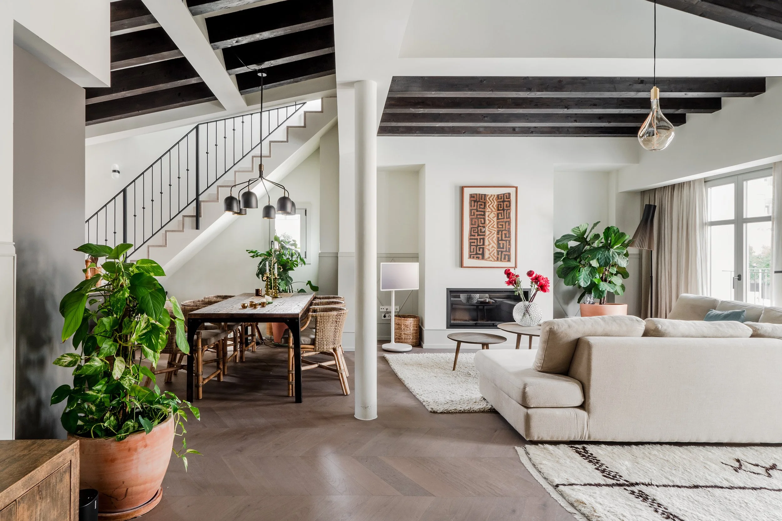 A modern living room with a dining area, featuring dark wooden beams on the ceiling, white walls, large windows with curtains, a beige sofa, a wooden dining table with chairs, and indoor plants, including a large potted plant in the foreground.