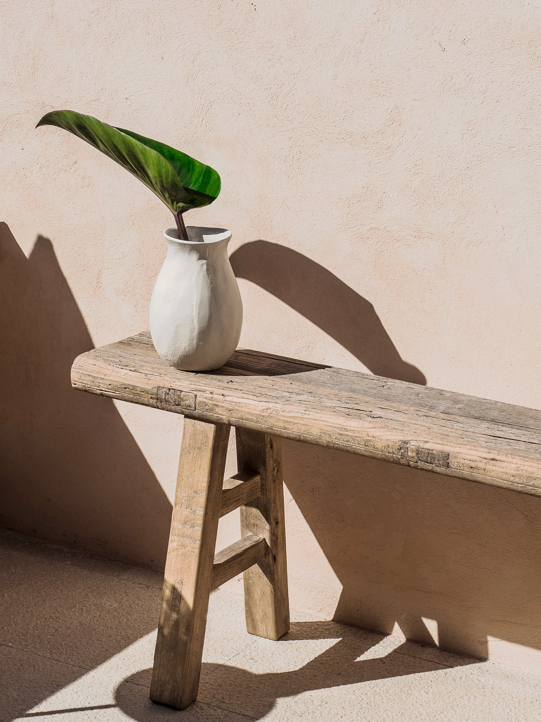 A white ceramic vase with a single green leaf on a wooden bench against a pink textured wall, casting shadows.
