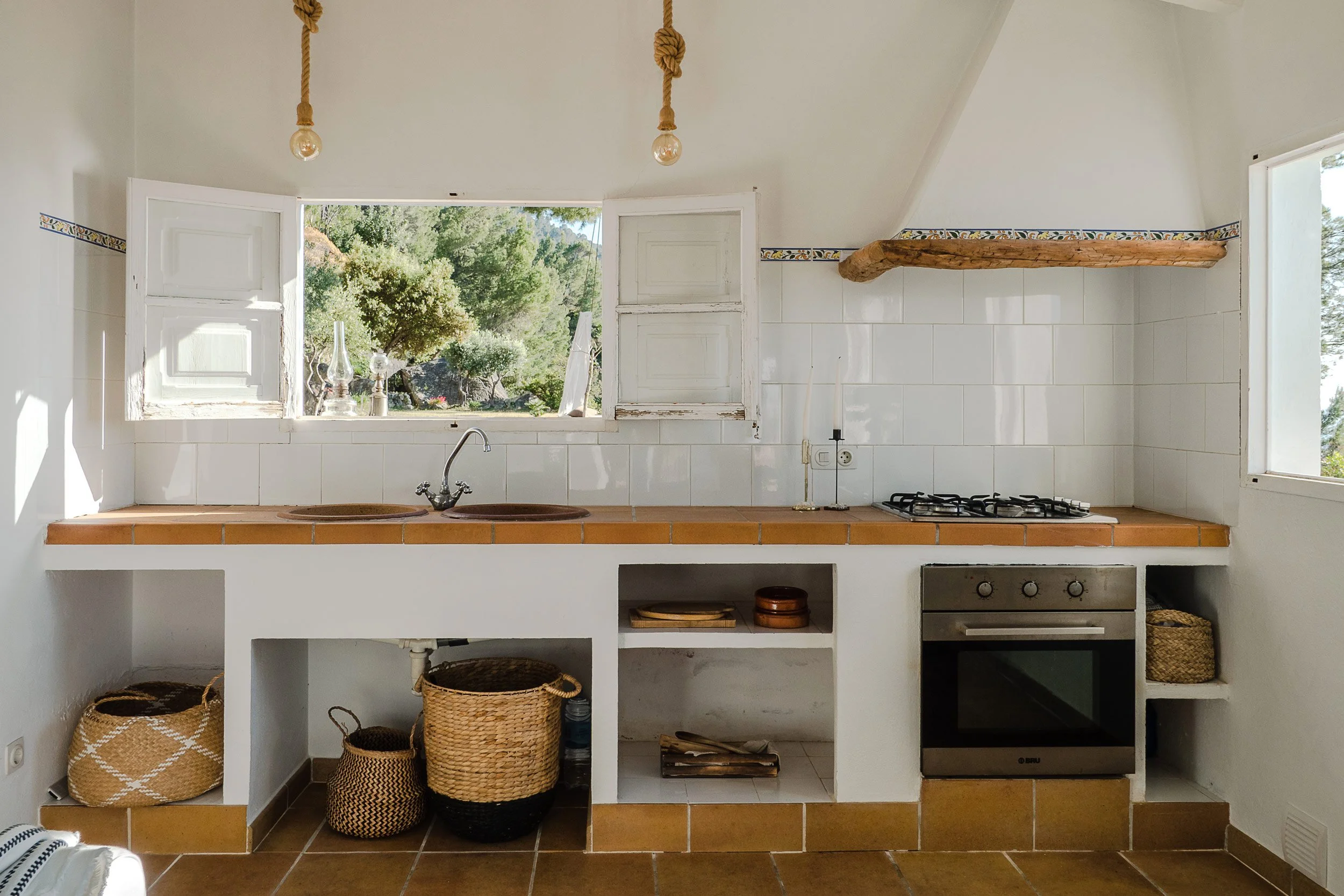 A kitchen with open white wooden shutters, a stainless steel sink, and a wooden shelf. There are woven baskets and pots below the counter, a gas stove, and a window showing greenery outside.