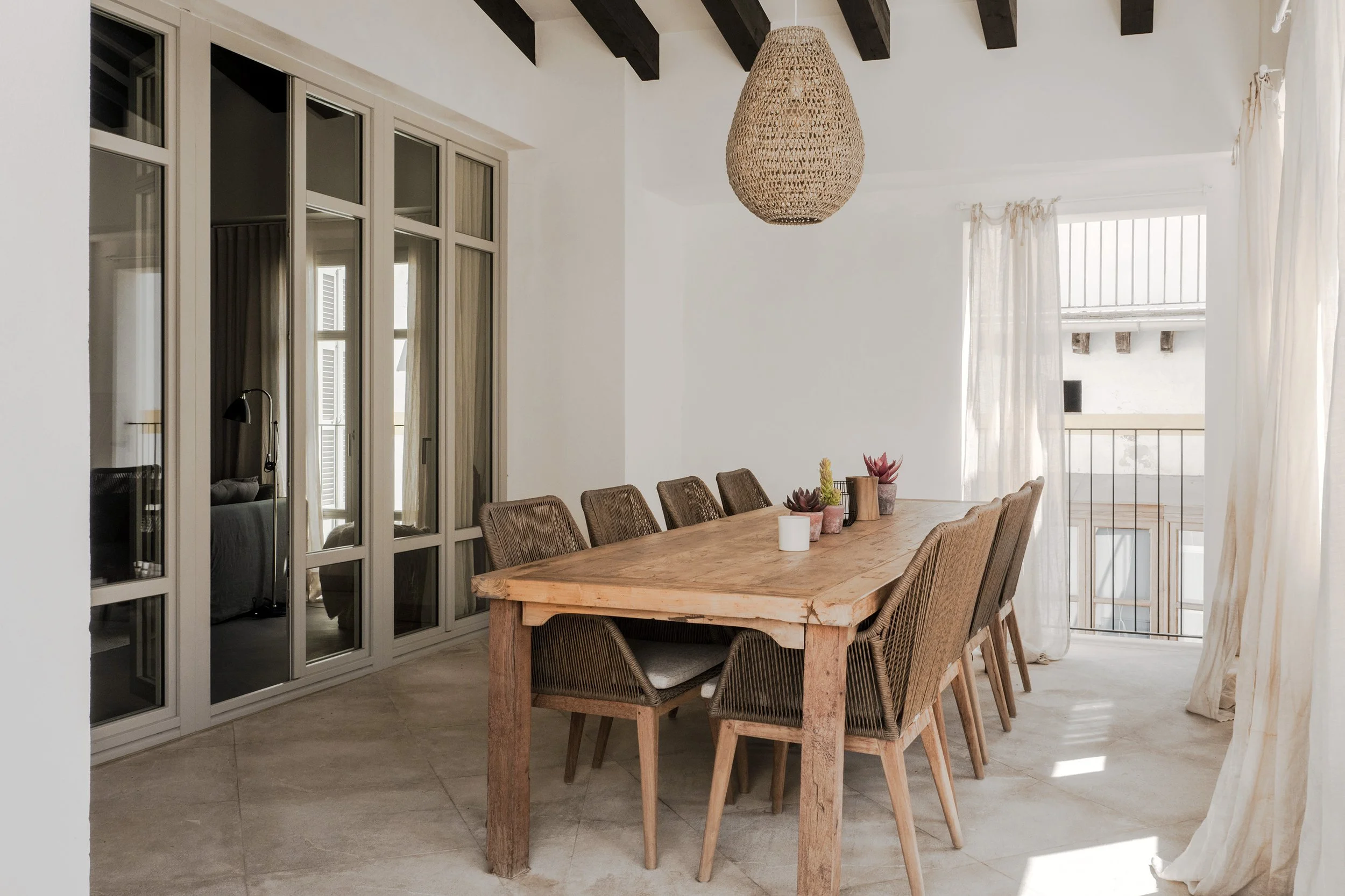 Dining room with a wooden table, wicker chairs, and a woven pendant light fixture, with large windows and cream curtains.