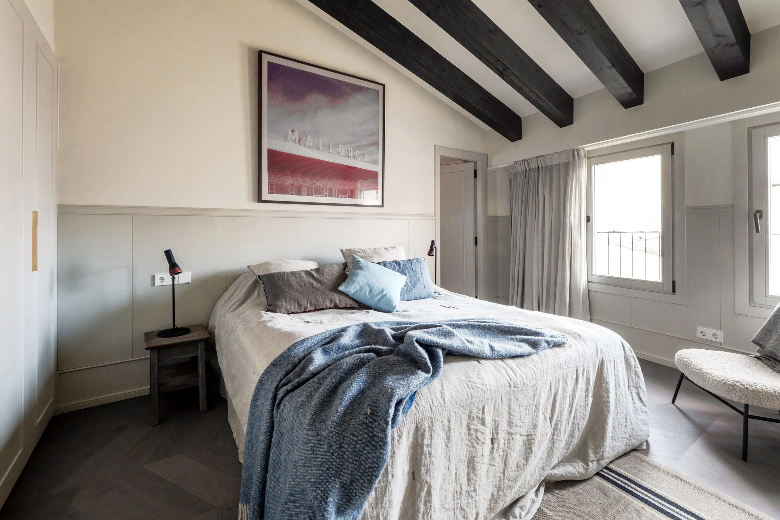 A cozy bedroom with a large bed covered in neutral bedding and a blue throw blanket. The bed has multiple pillows, including a beige pillow and a blue decorative one. On each side of the bed, there are small nightstands with black reading lamps. The 