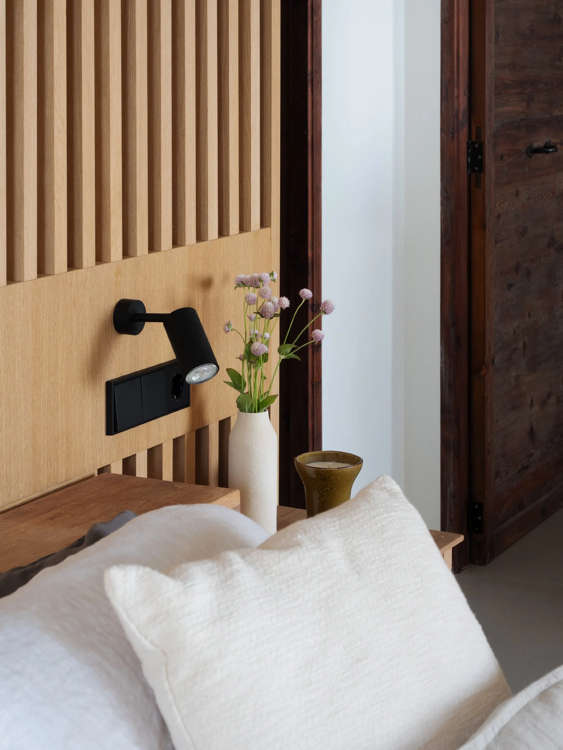 Close-up of a bed with a white quilt, a bedside table, a vase with pink flowers, and a wall-mounted reading light in a bedroom.