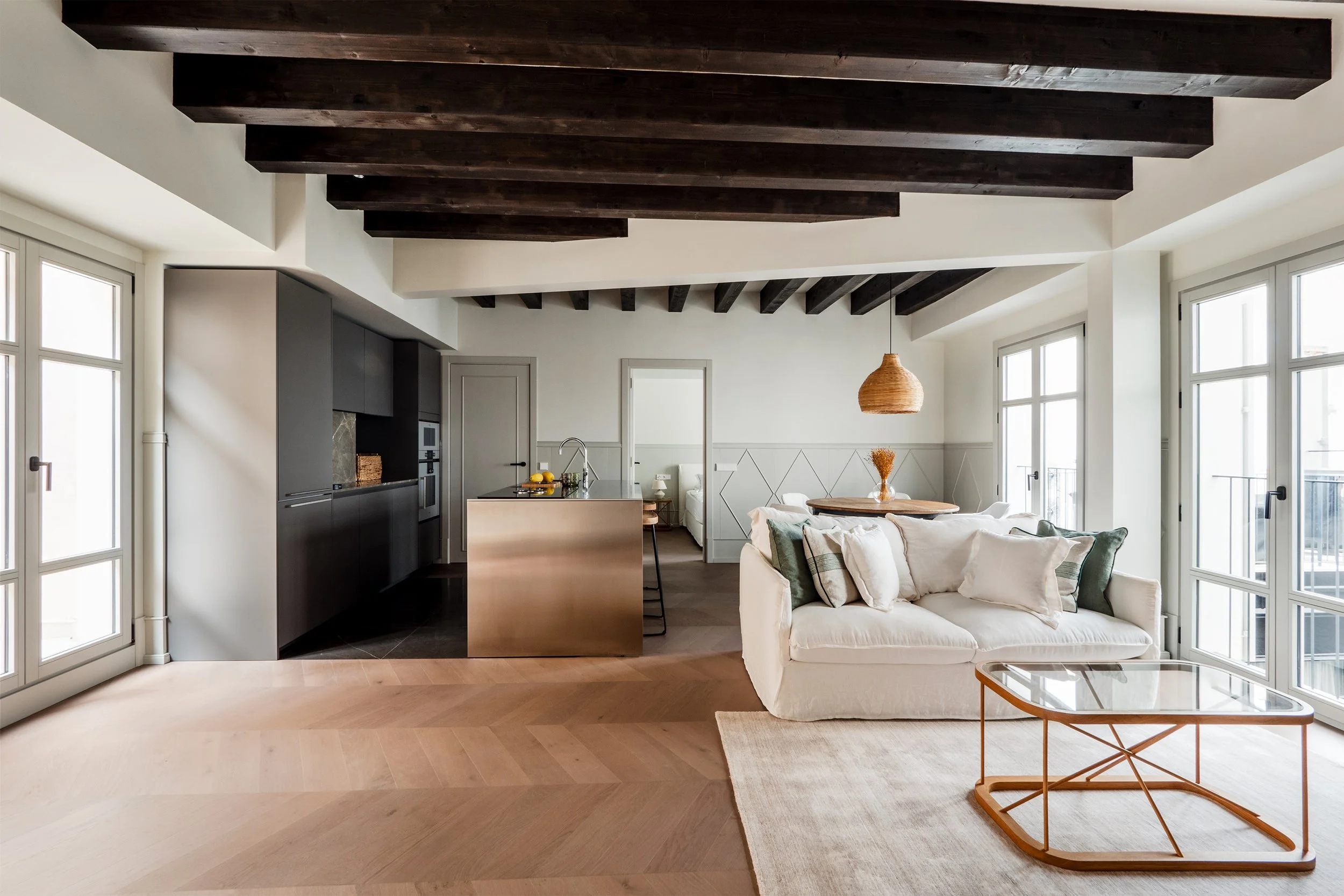 Open-concept living area with white walls, large windows, dark wooden ceiling beams, a white sofa with green and beige pillows, a glass coffee table on a beige rug, a wooden round table with a vase of orange flowers, and a modern kitchen with black c