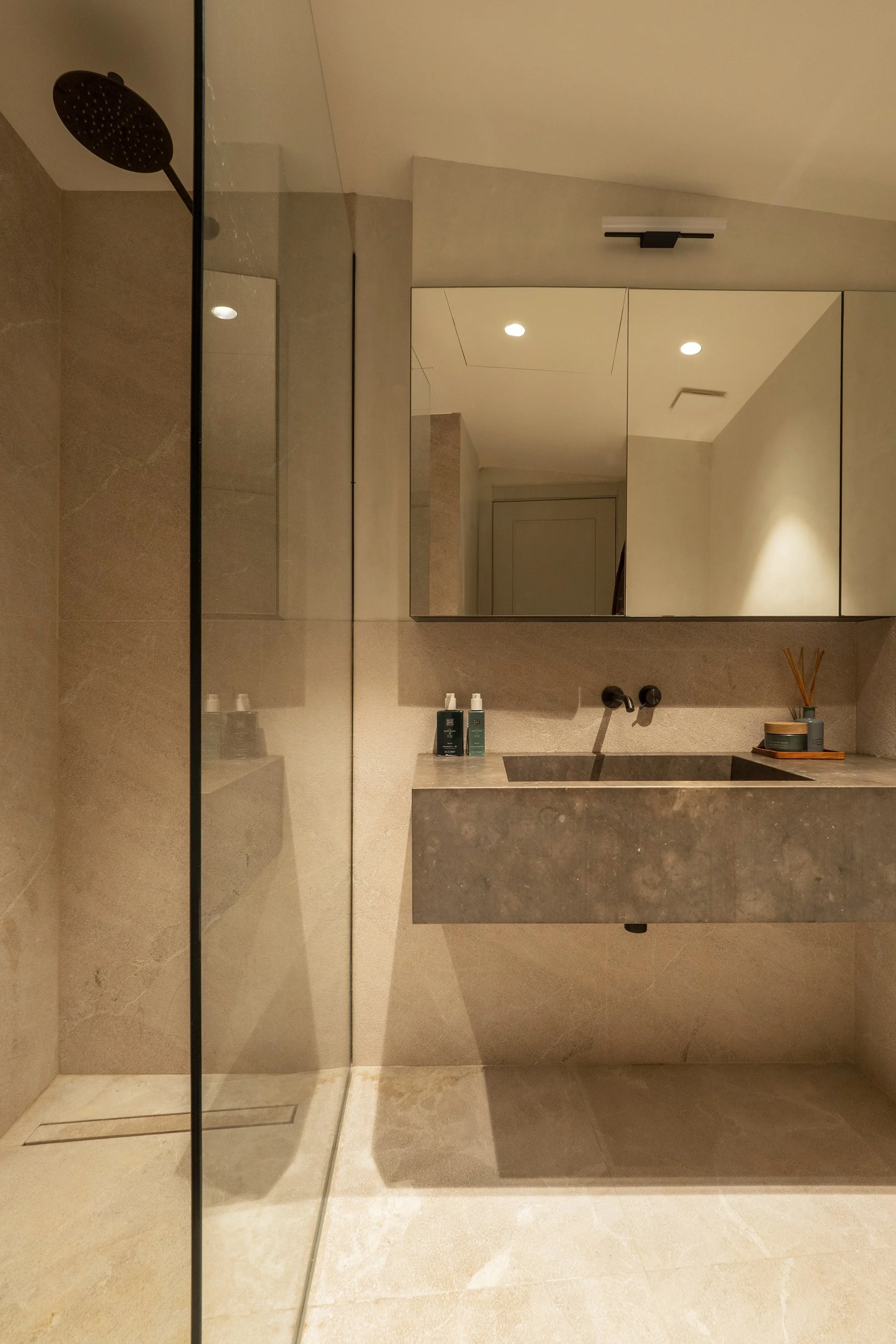 A modern bathroom with a walk-in shower, a large mirror, a stone vanity with a sink, and minimal decor. The shower has a rain shower head and a glass partition. The bathroom is illuminated by recessed ceiling lights.