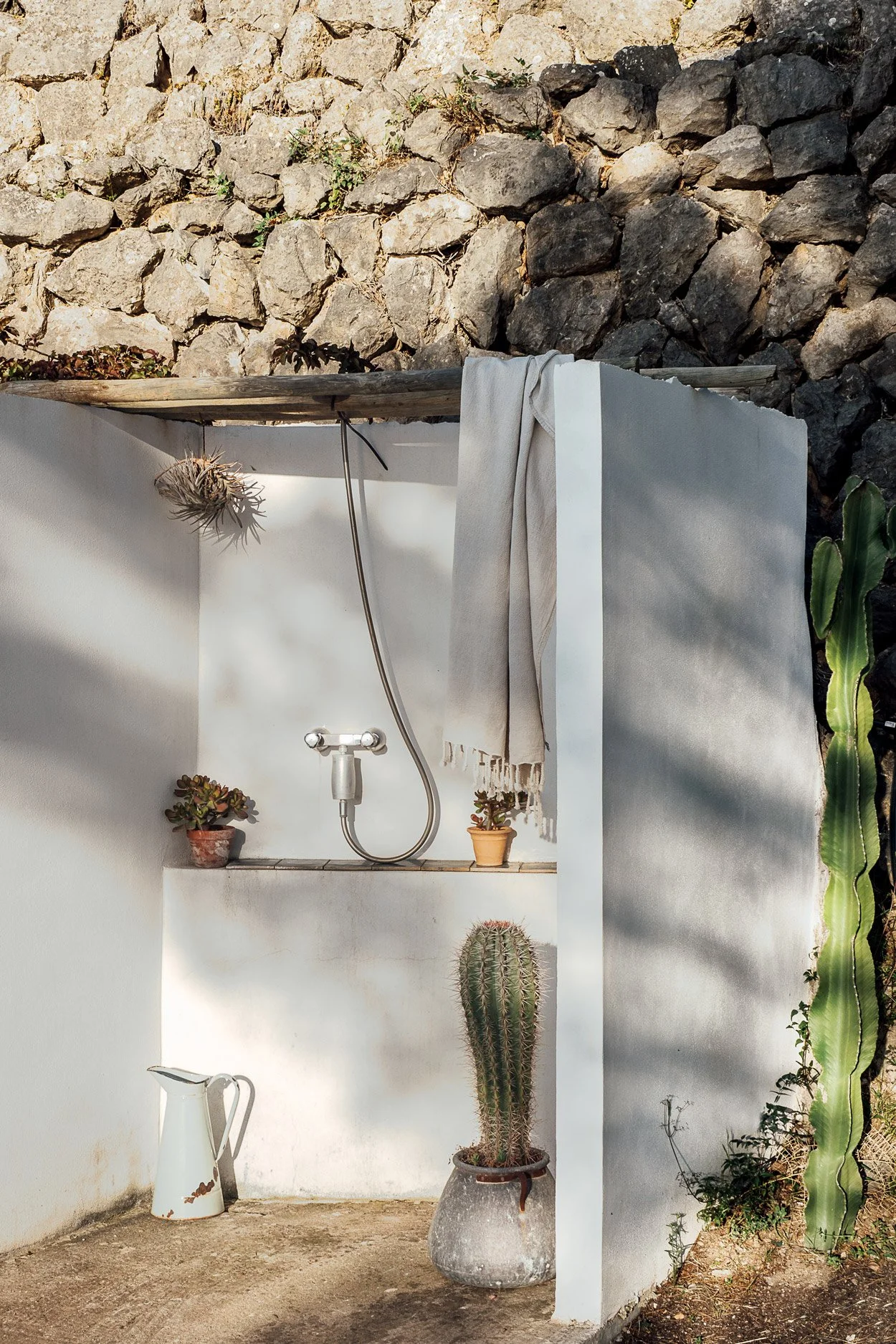An outdoor shower area with a white wall, a hanging towel, small potted plants, a tall cactus in a pot, a white pitcher, and a stone wall backdrop.