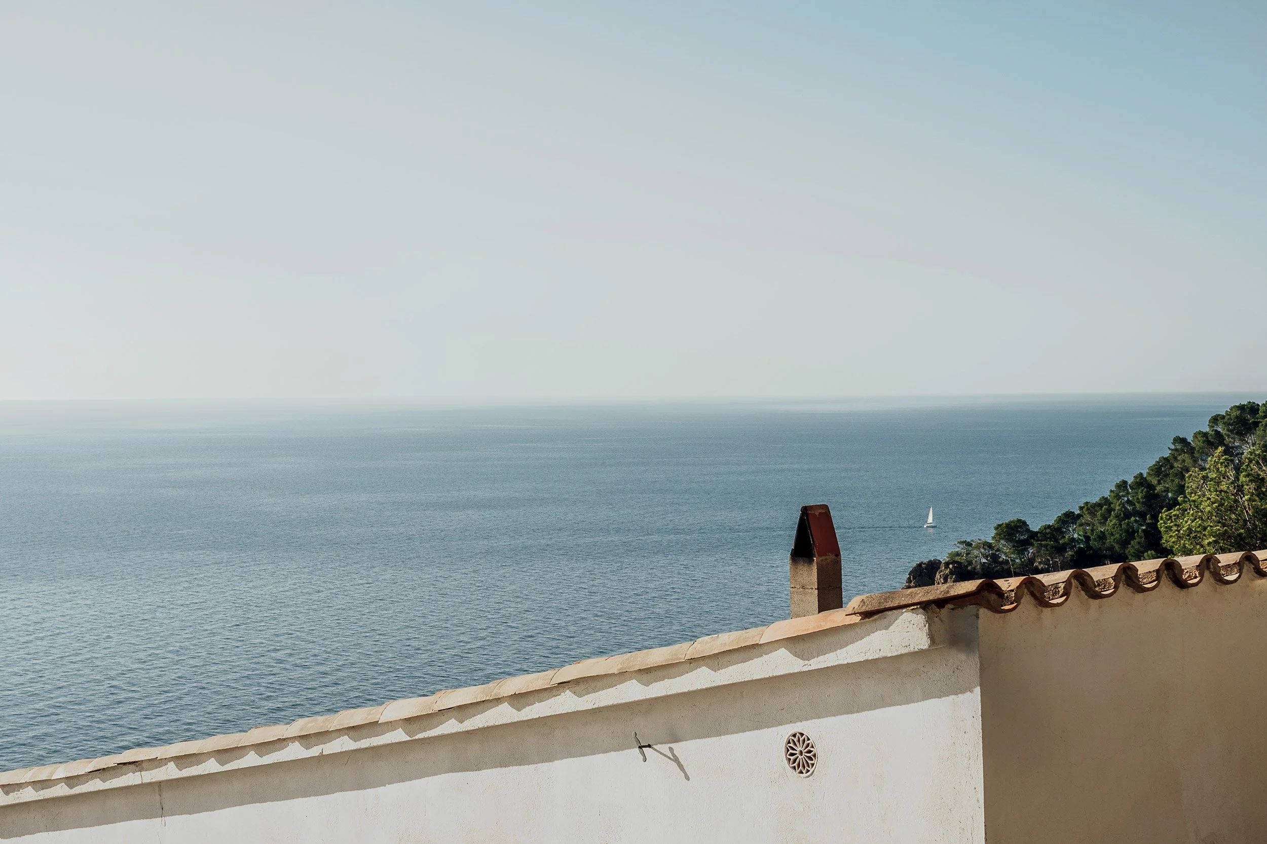 View of the ocean with a sailboat in the distance, seen from a balcony with a white stucco wall and a tiled roof, with trees on the right side.