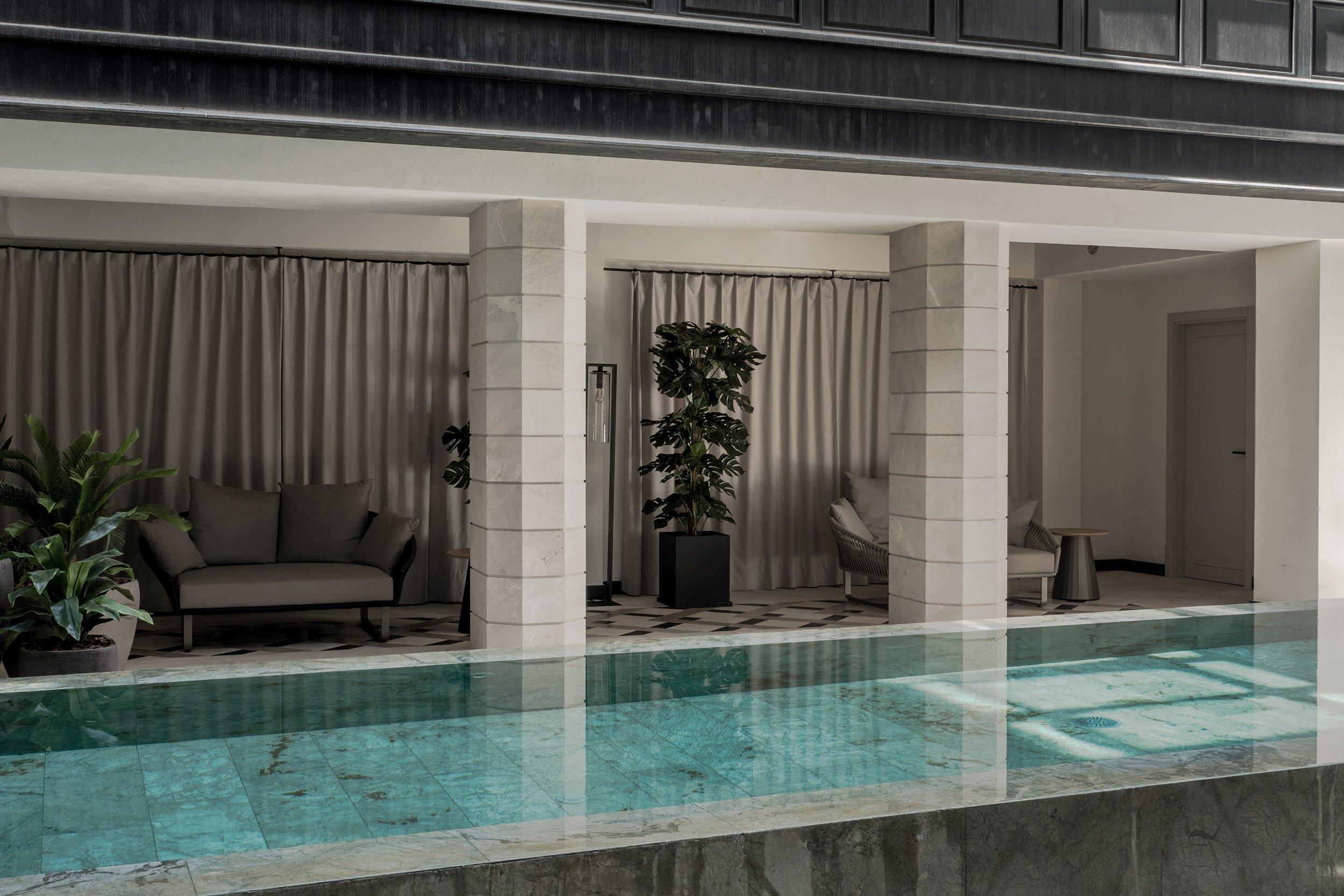 Indoor lounge area with beige couches, potted plants, beige curtains, and a tiled floor, adjacent to a swimming pool with green and blue tiles.