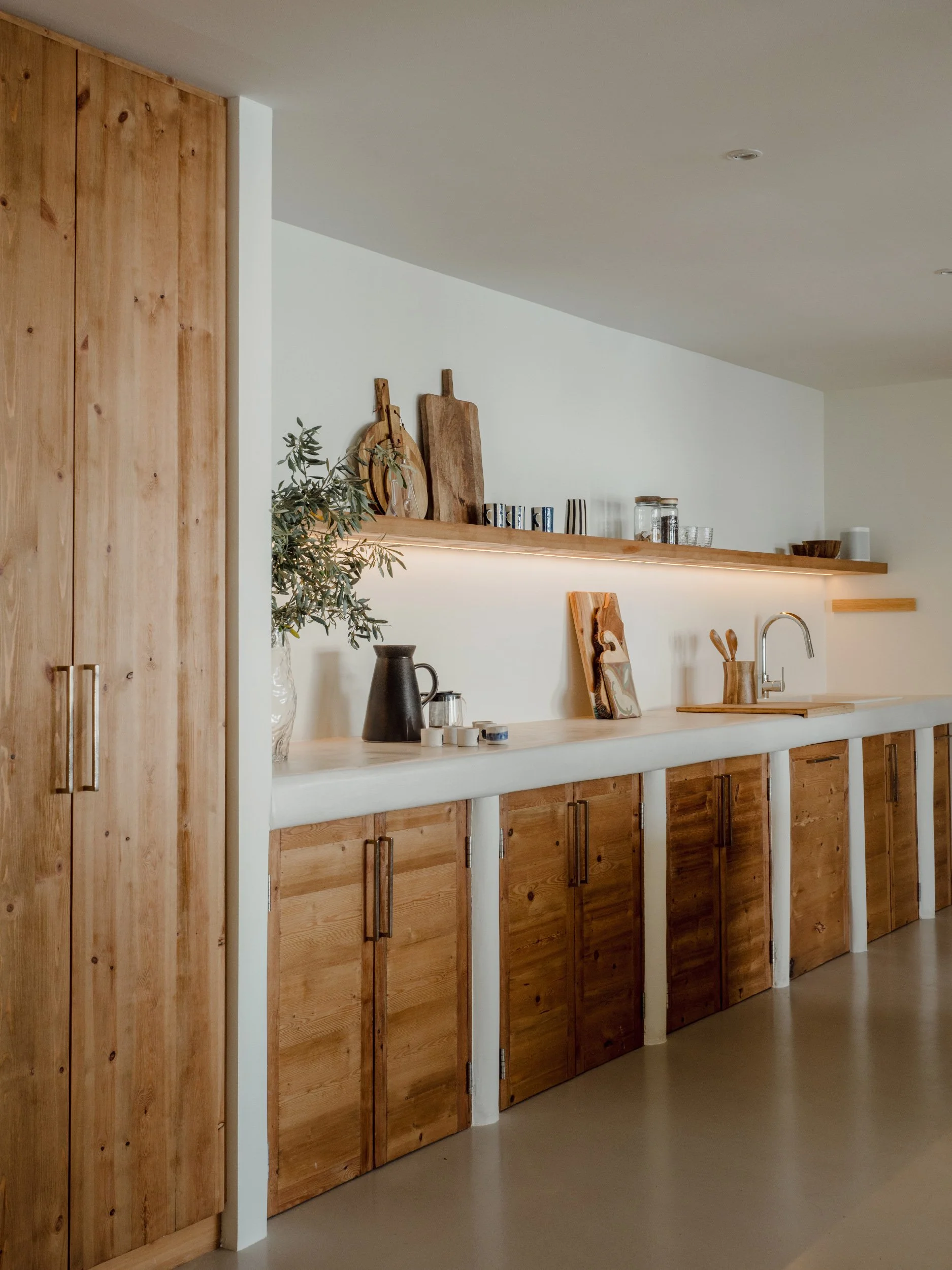 Wooden cabinets lined up in a kitchen with a white countertop, a black pitcher, and a small plant in a glass vase, with cutting boards and jars on the shelves above.