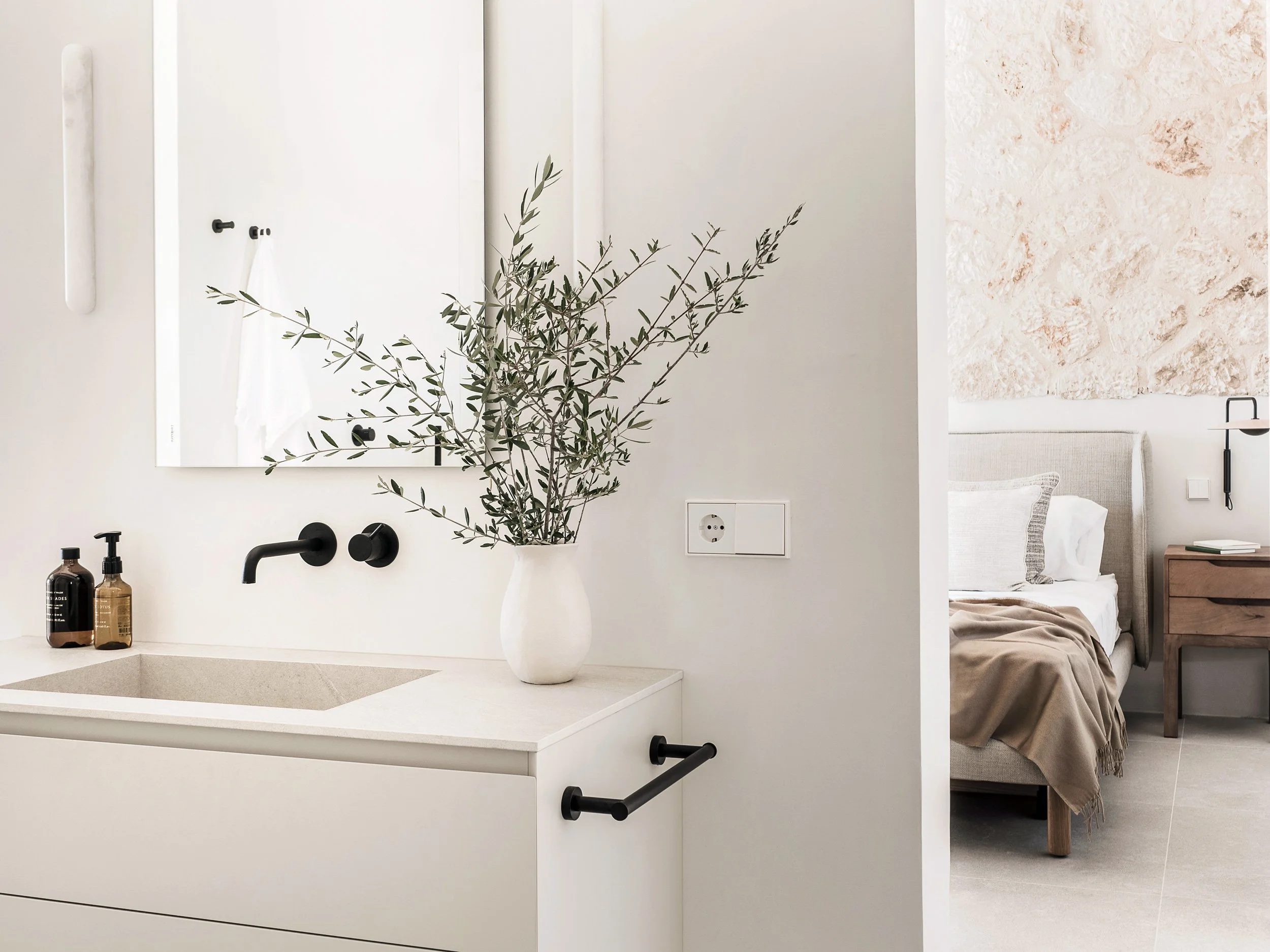 Modern minimalist kitchen with white cabinetry, black fixtures, a botanical vase, and a bathroom with a large mirror reflected in the background.