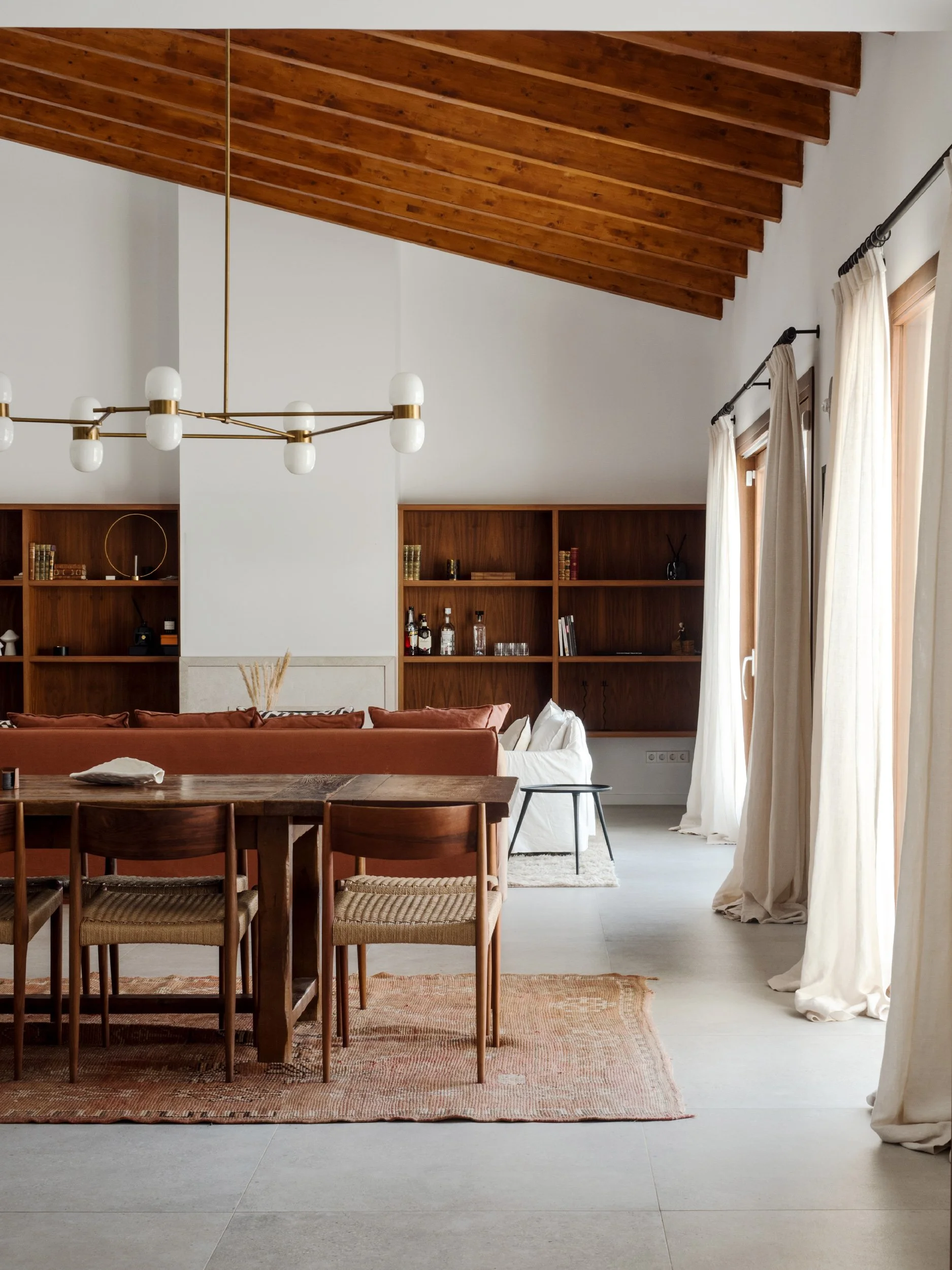A living room with a wooden ceiling featuring exposed beams, a modern chandelier with round white bulbs, a wooden dining table with matching chairs on a patterned rug, a white couch with rust-colored pillows, and built-in wooden bookshelves with books and decorative items. Large cream curtains cover the windows.