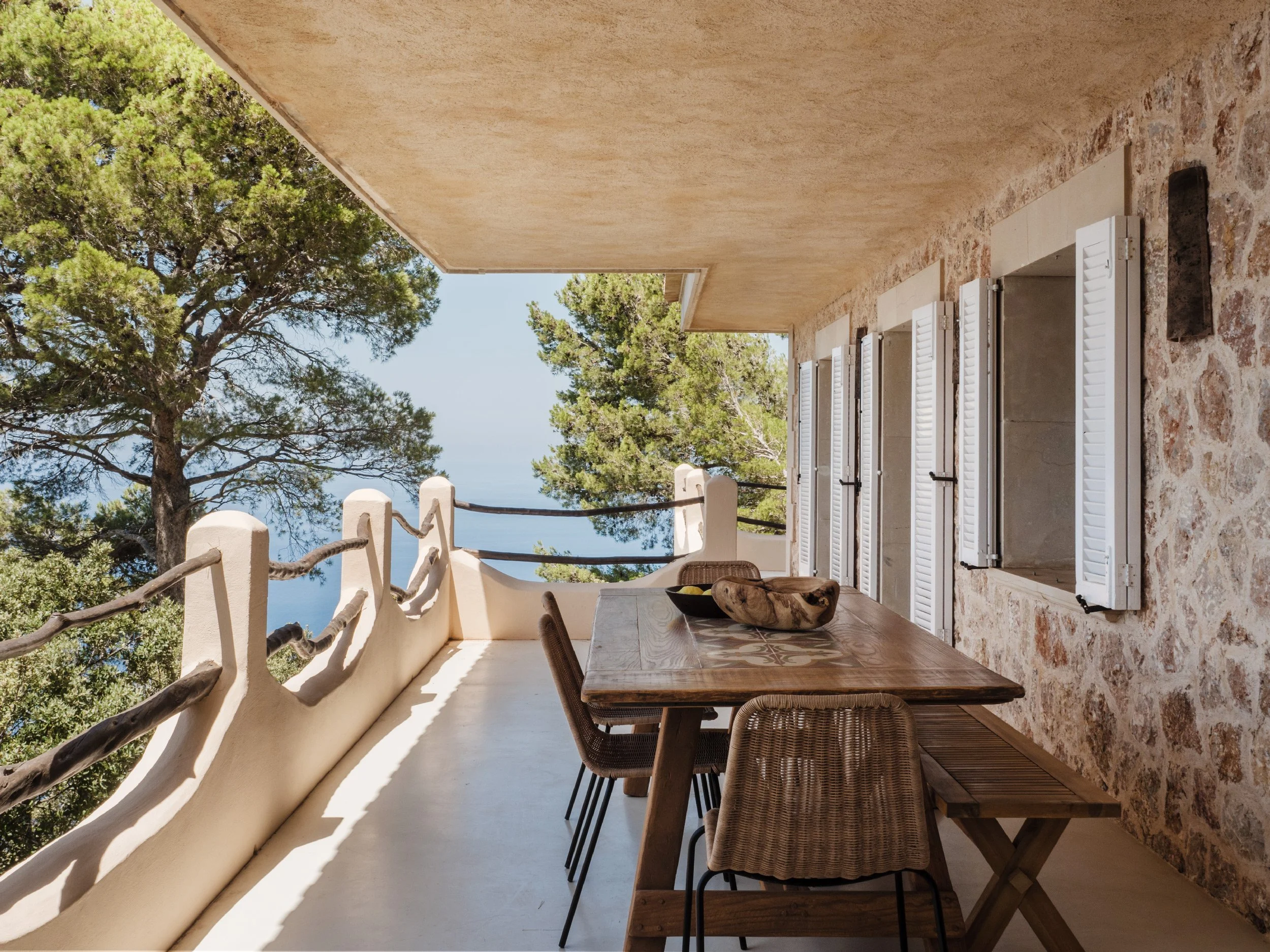 Balcony with a rustic wooden table and chairs, white shutters on a window, a stone wall, and green trees in the background under a clear sky.
