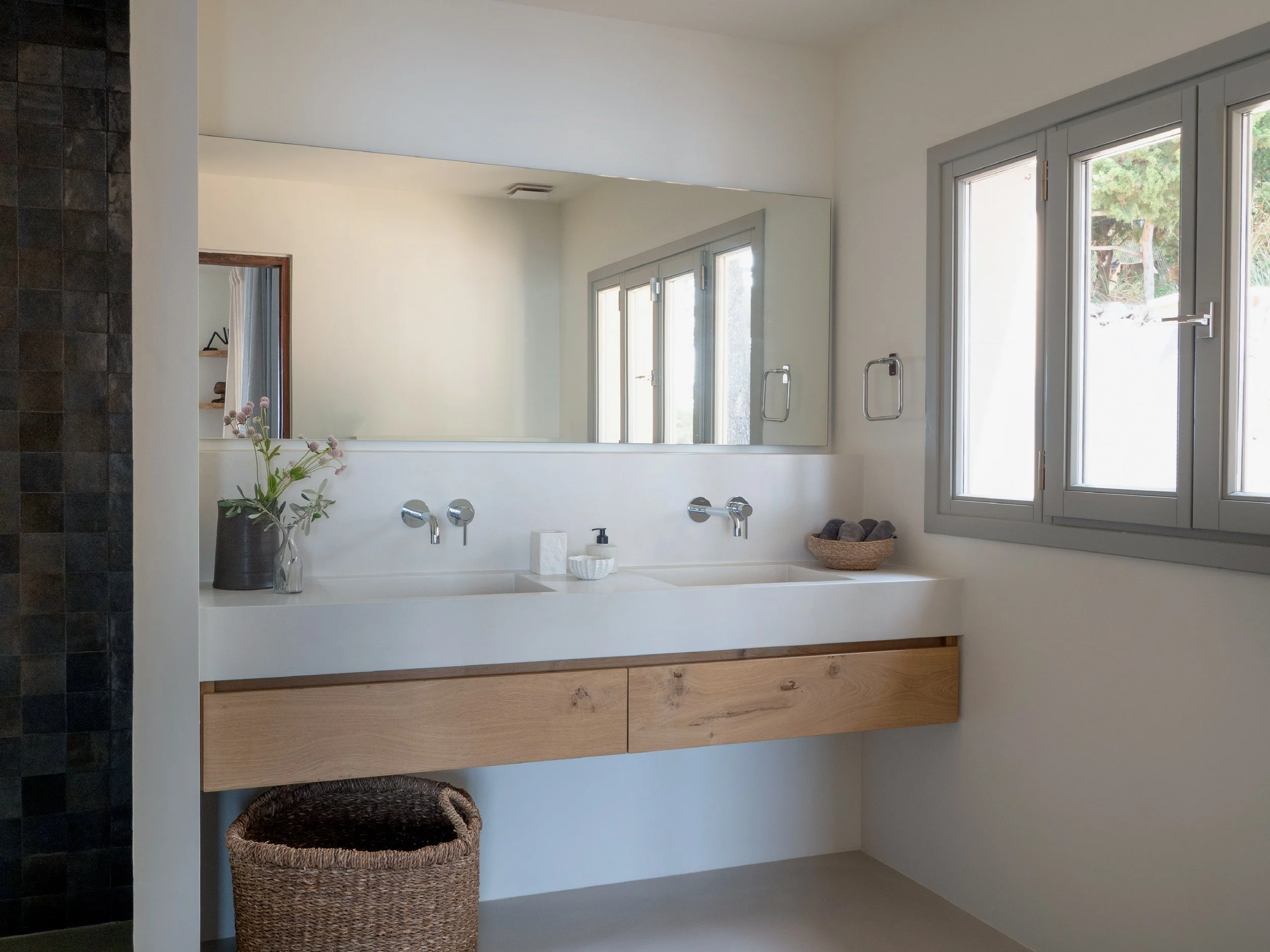 Modern bathroom with a large mirror, white sink, black vase with flowers, wicker laundry basket, and large window with scenery outside.