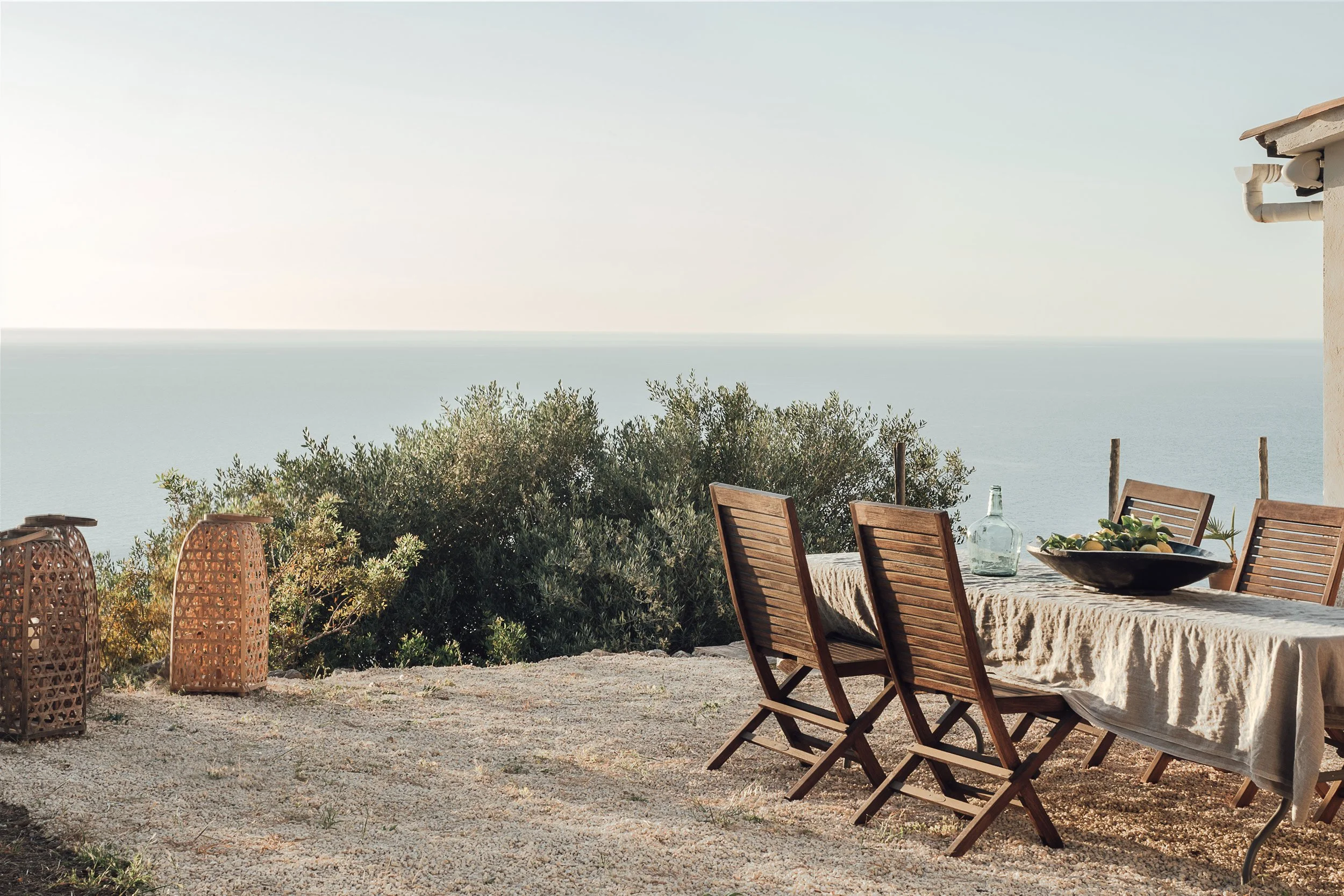 An outdoor dining area with wooden chairs around a table covered with a cloth, set against a backdrop of shrubbery and an ocean view, with a clear sky.