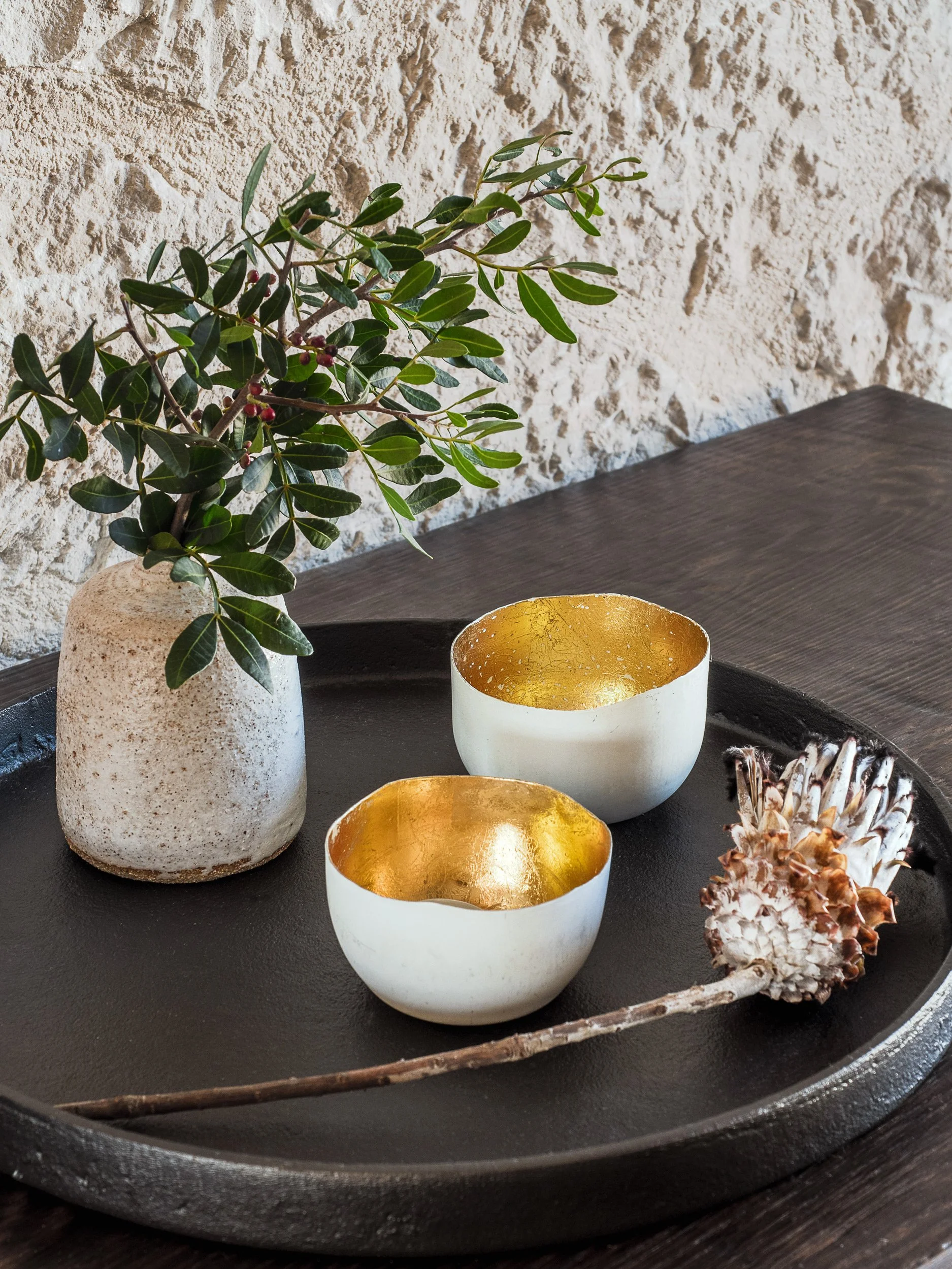 Decorative display with a green leafy plant in a textured beige vase, two white bowls with gold interiors, and a dried pine cone on a black tray.