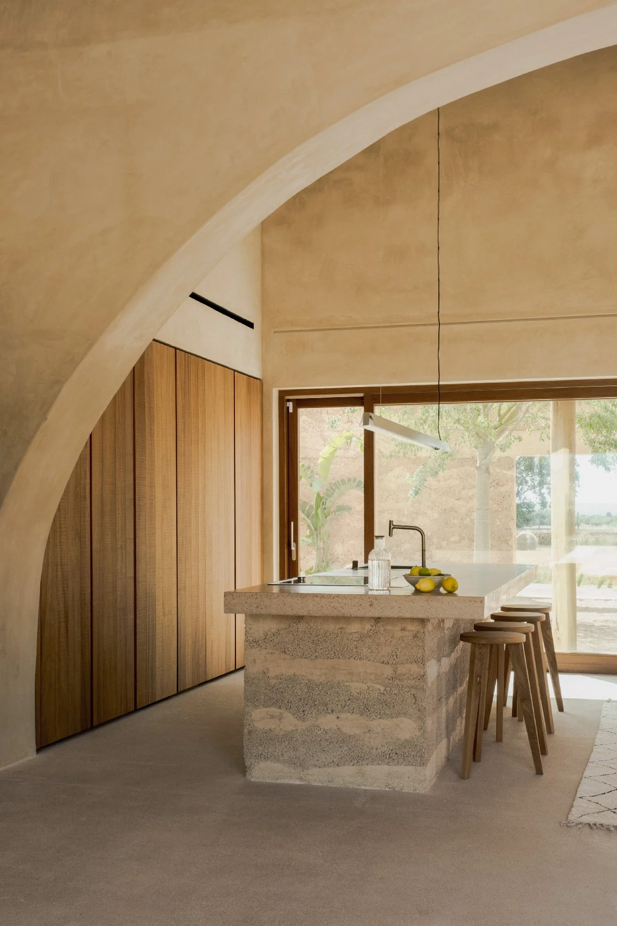 A modern kitchen with a stone island, wooden cabinets, a large window, and bar stools.