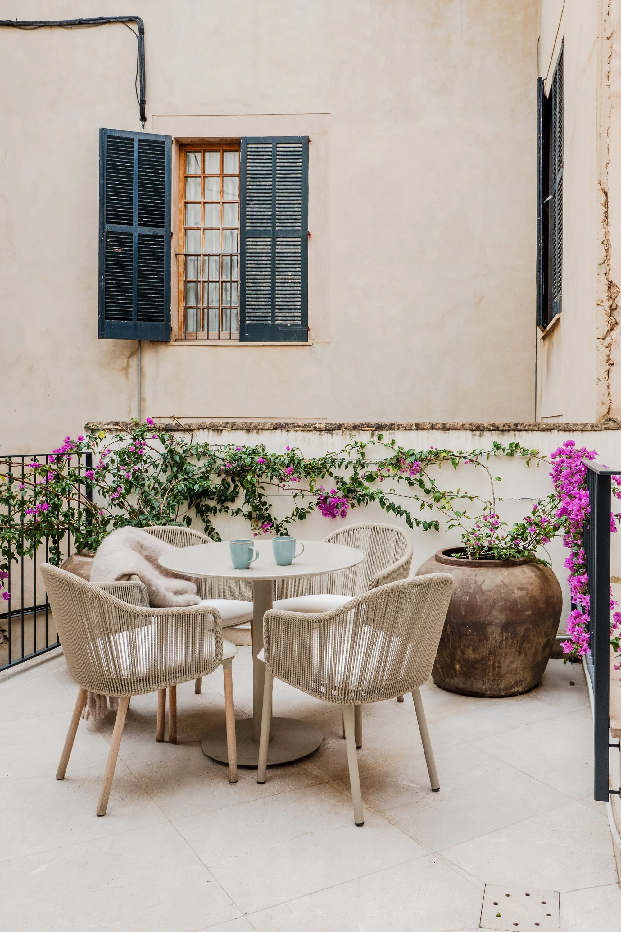Outdoor patio with a round table, six beige chair, two light blue cups, large flower pot, green vine with pink flowers, cream-colored walls, closed window with wooden frame and blue shutters.