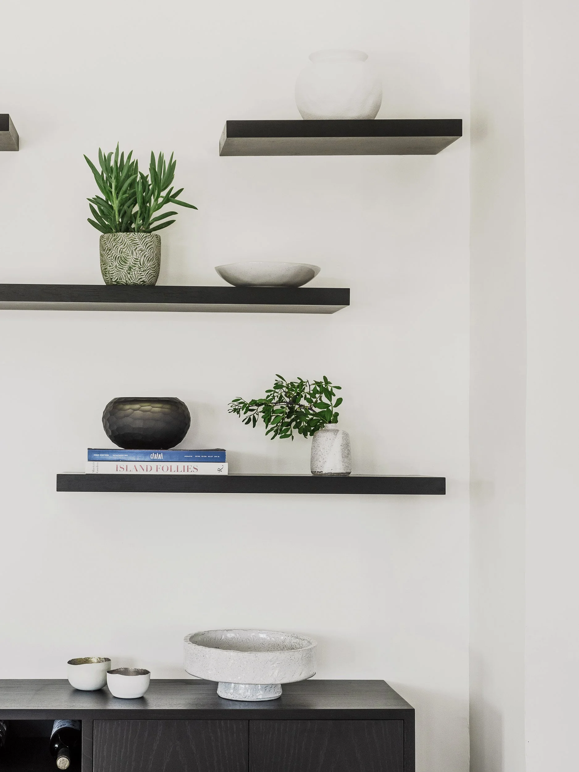 Interior of a modern living space with black floating shelves holding decorative items, including a white pot with a green plant, a shallow bowl, and a white ceramic vase with greenery. A black sideboard below displays two white bowls, a large textur