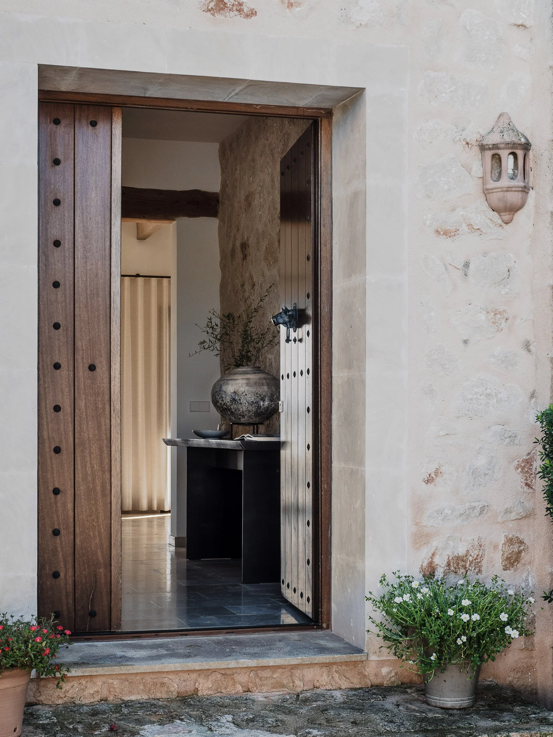 A doorway with a wooden frame and a black door handle, set in a textured beige stone wall, with potted plants on either side.