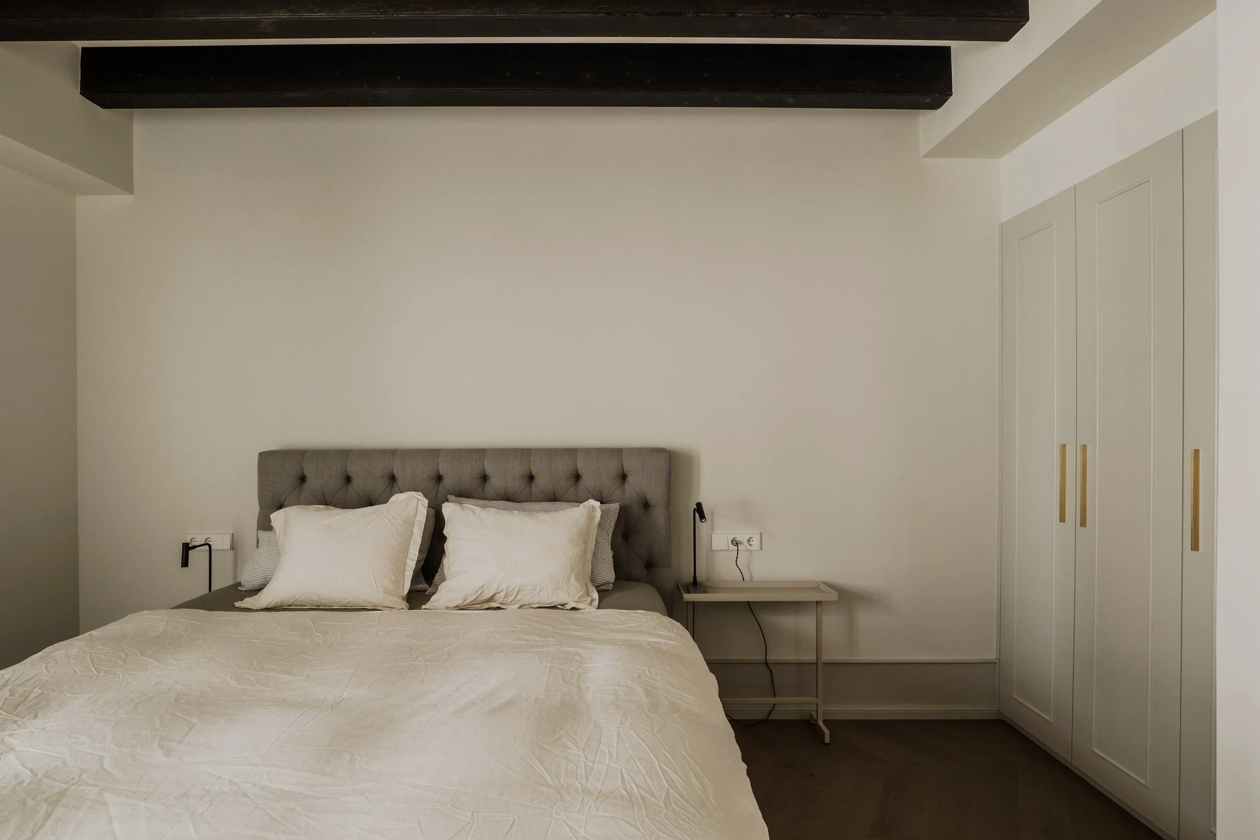 A bedroom with a gray upholstered headboard, white bedding, and pillows. A small white bedside table on the right with a black lamp and a nightstand on the left with a black lamp. White walls with a power outlet and cable on the wall next to the bed.