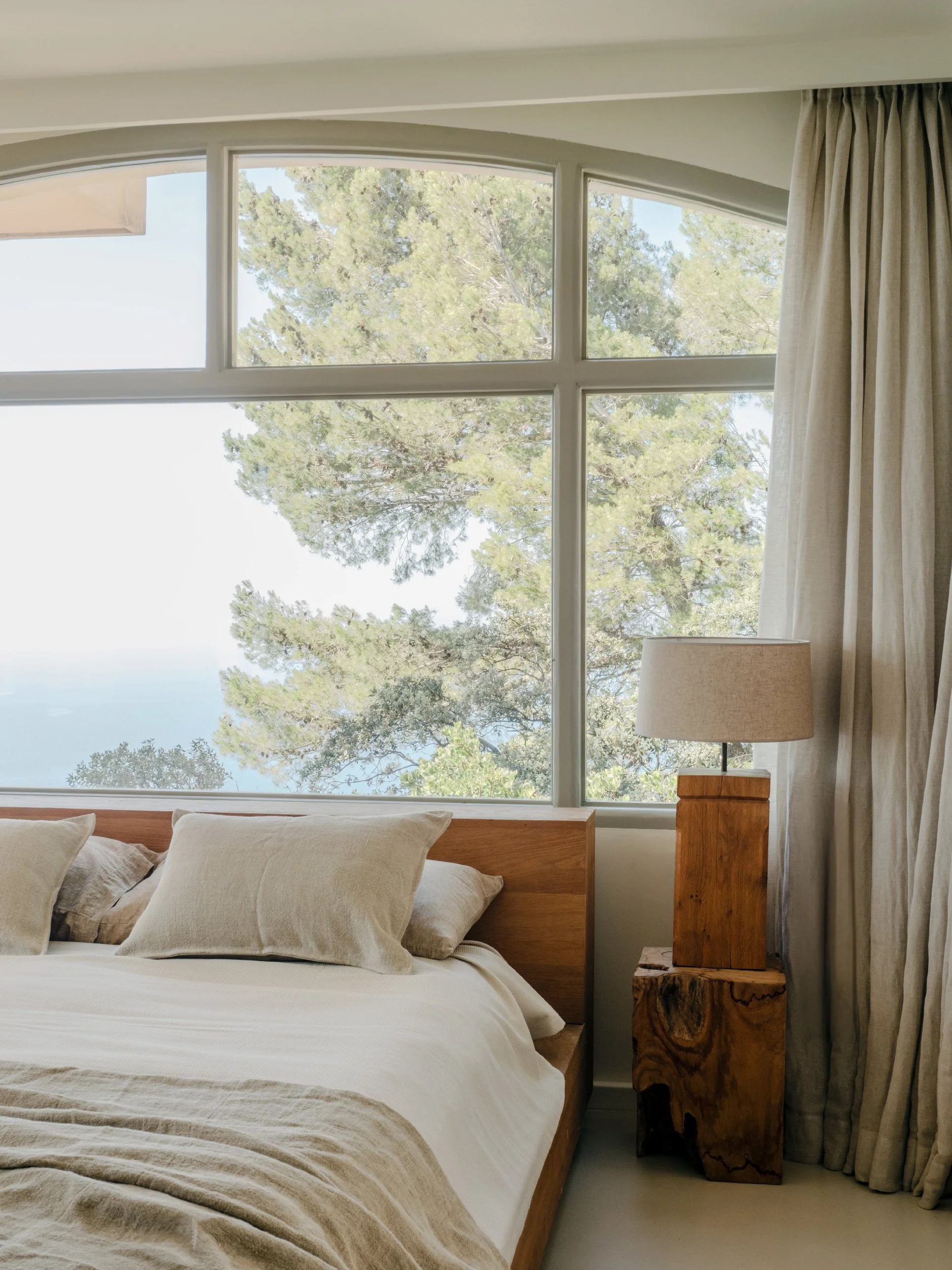 A cozy bedroom with a large window showing leafy trees outside. The bed has beige linens and pillows. There is a wooden side table with a unique natural wood finish holding a beige lampshade.