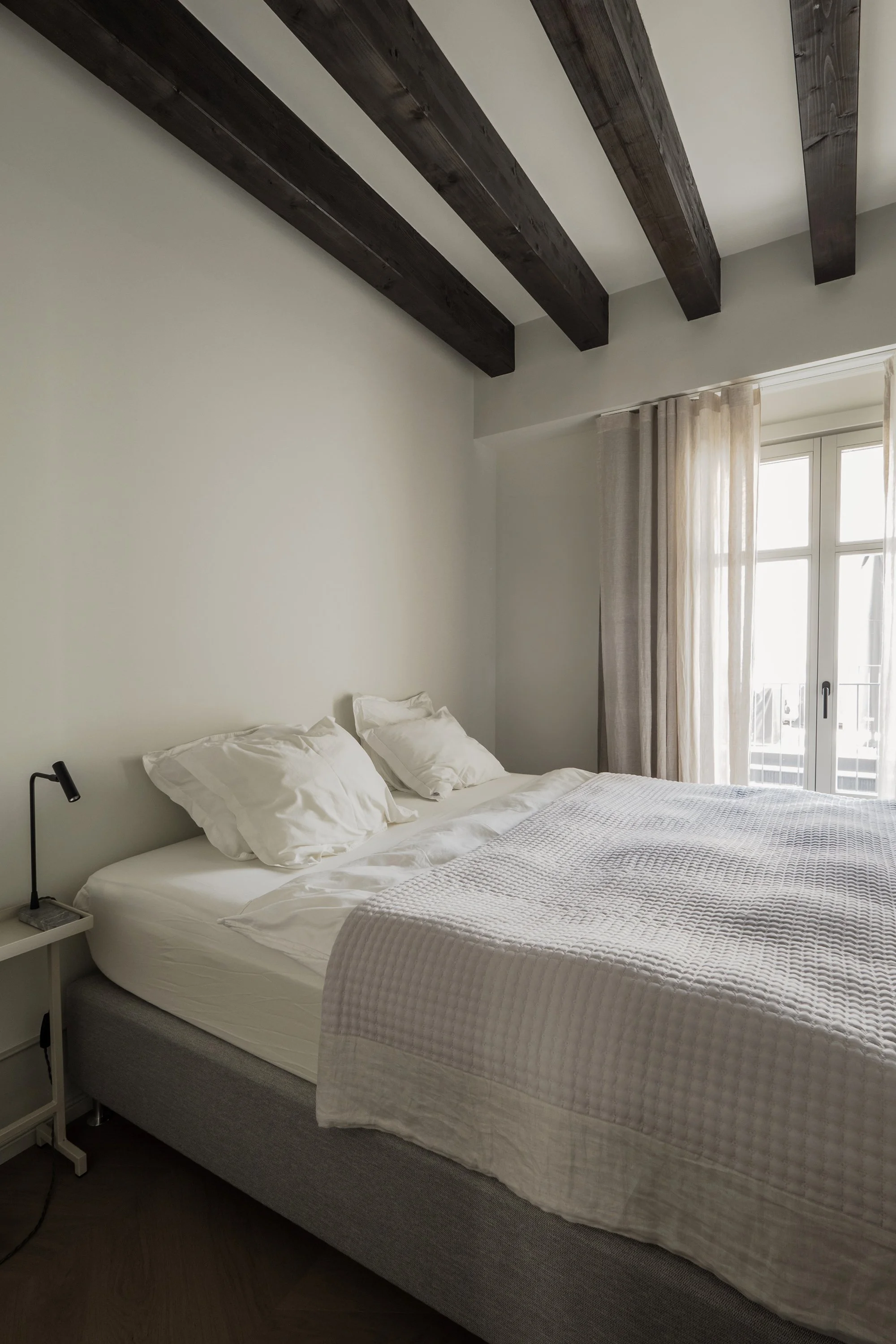 A neatly made bed with white bedding and four pillows, situated in a bright room with a window and beige curtains. The ceiling has dark wooden beams.