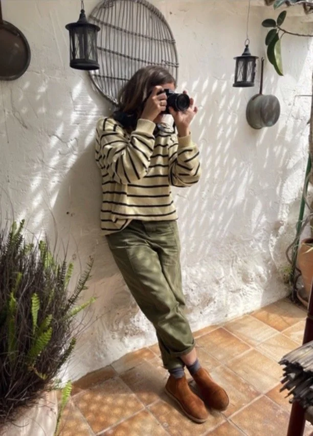 Person with shoulder-length hair and striped sweater taking a photo with a camera, standing against a white textured wall with hanging lanterns and plants.