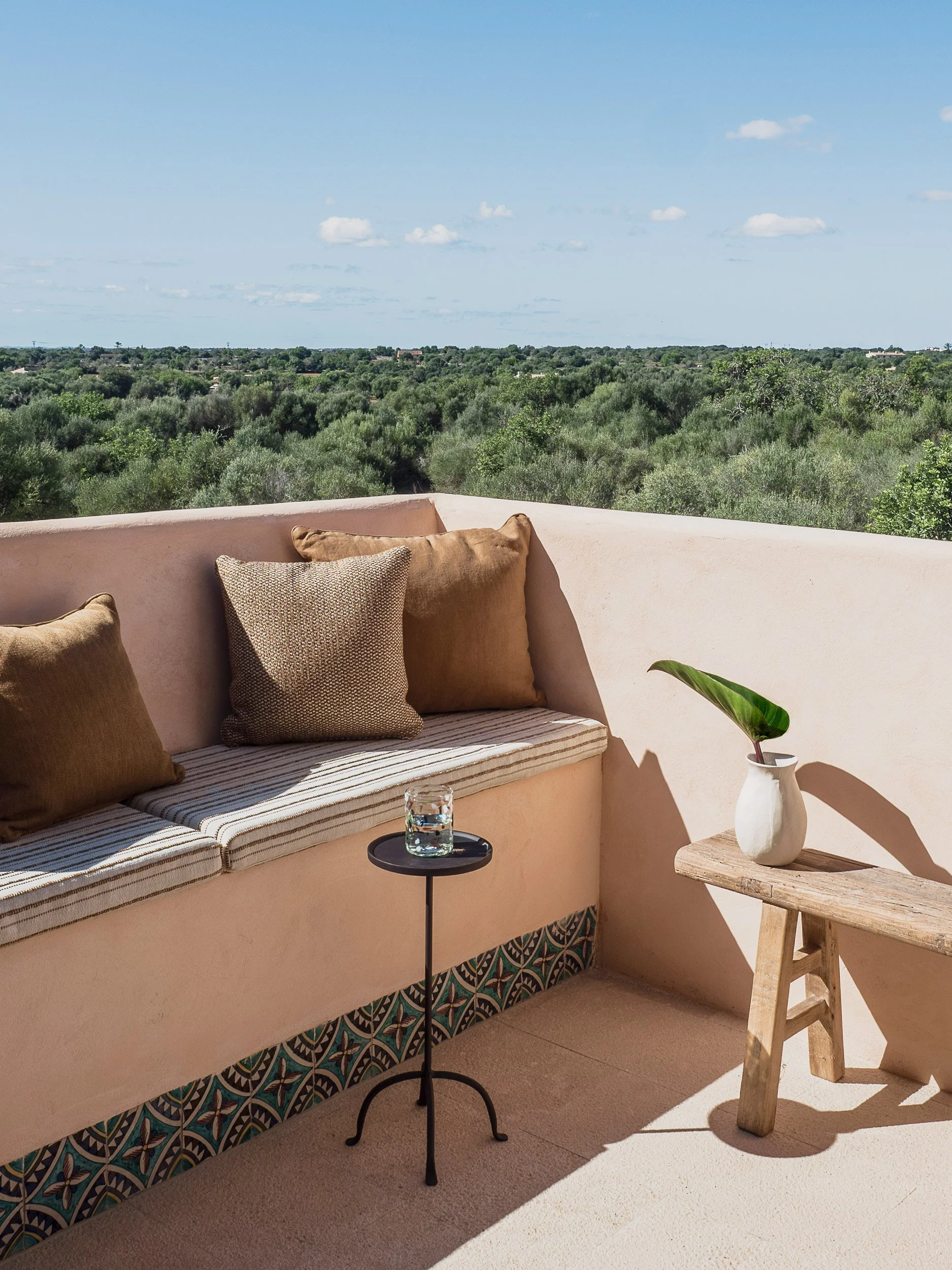 A rooftop patio with pastel pink walls and a built-in cushioned seating area decorated with beige throw pillows. There is a small round black side table with a glass of water, a wooden stool with a decorative vase containing a large green leaf, and a